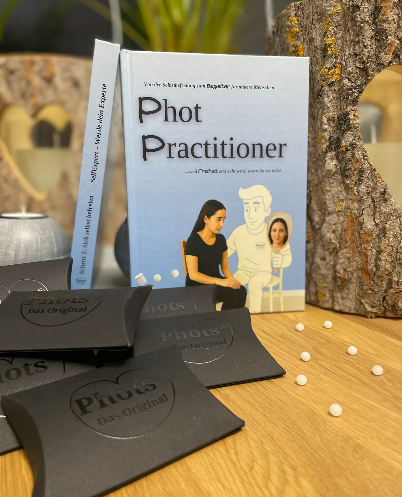 Buch mit dem Titel 'Phot Practitioner' mit Illustration einer Frau und einer Cartoon-Figur, umgeben von schwarzen Verpackungen mit der Aufschrift 'Phots Das Original' auf einem Holztisch.