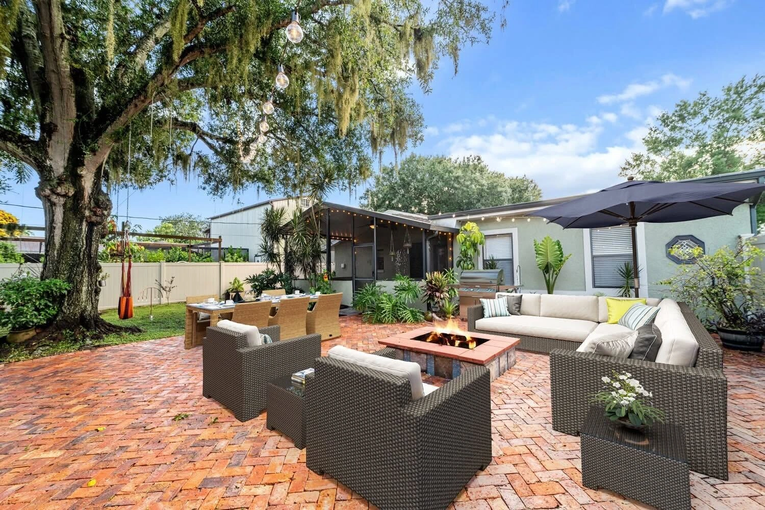 A cozy backyard with a brick patio features a fire pit surrounded by wicker chairs and a sectional sofa. 