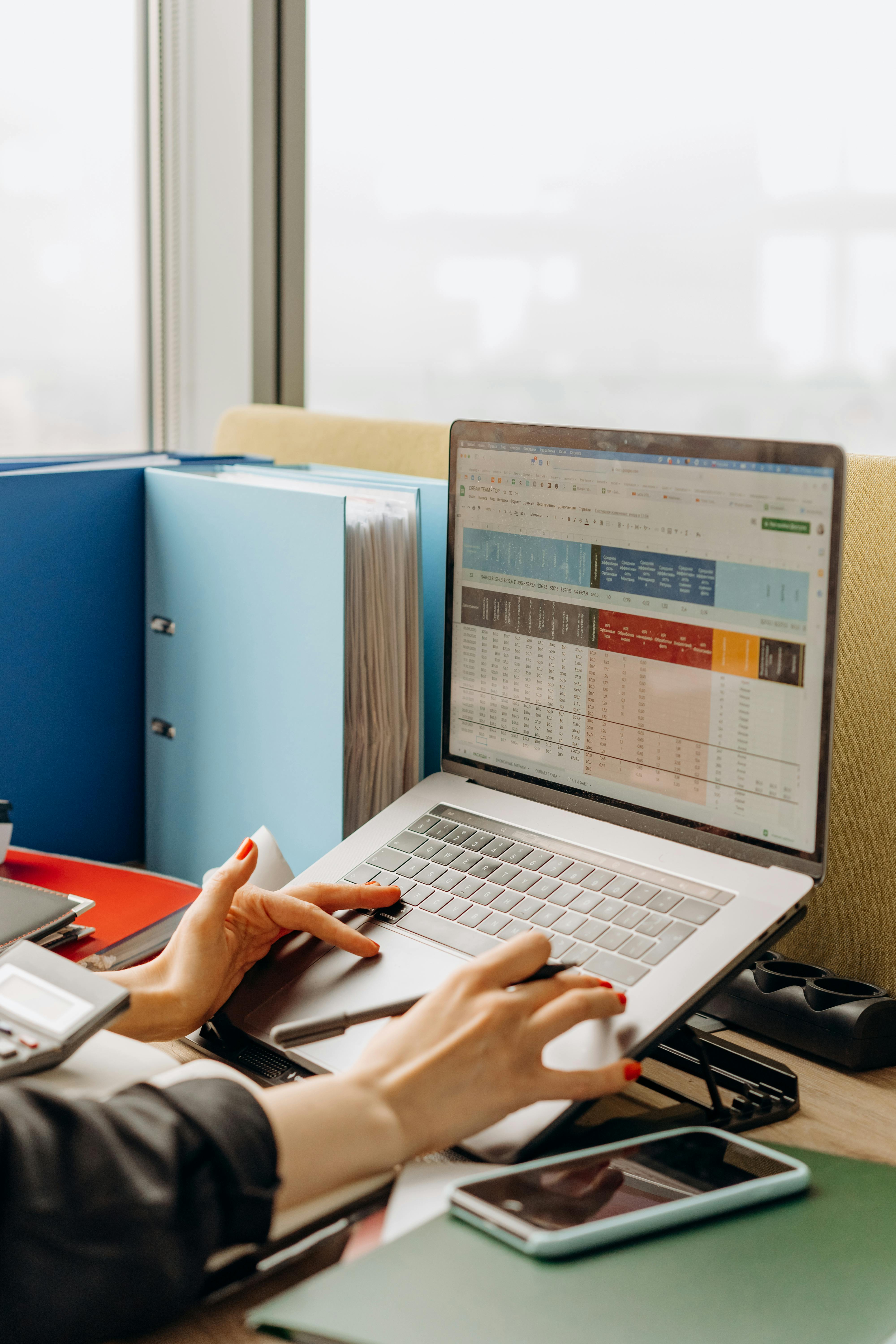 Person working on a laptop displaying a colorful spreadsheet, with folders, a smartphone, and office supplies on the desk.