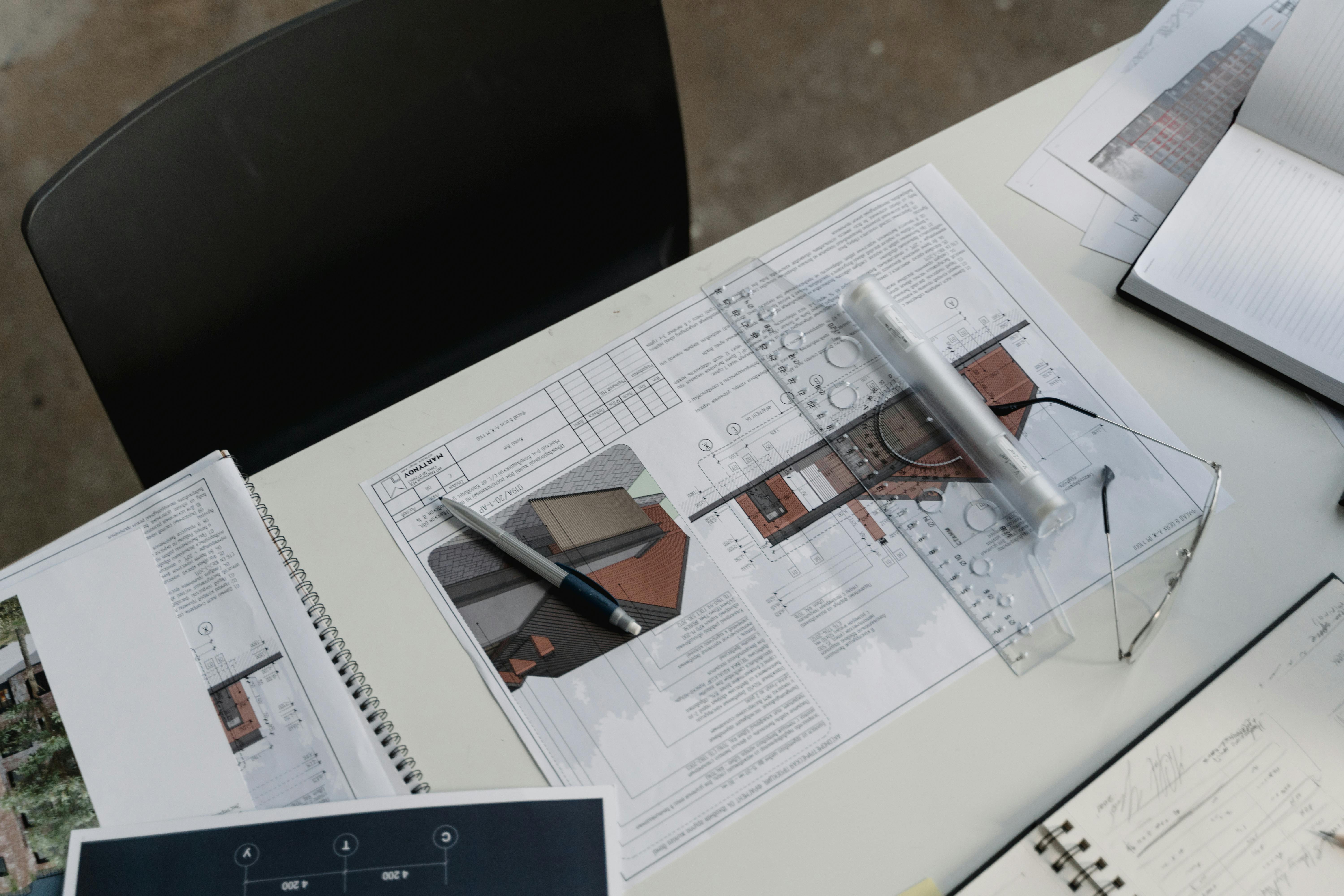 Architectural blueprints and sketches spread on a white desk with a pen, transparent ruler, and eyeglasses.