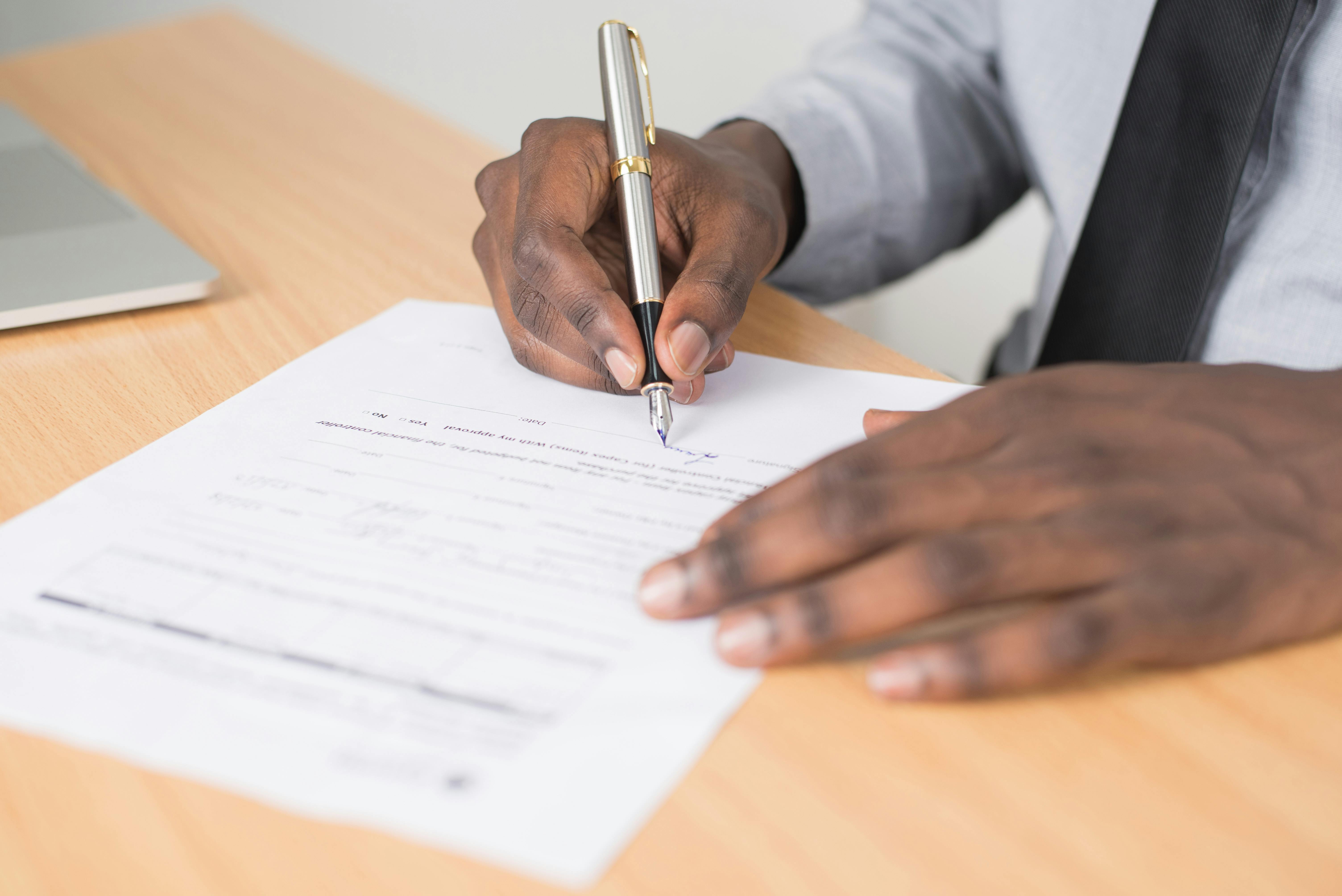 Person in business attire signing a document with a silver and gold pen on a wooden desk.