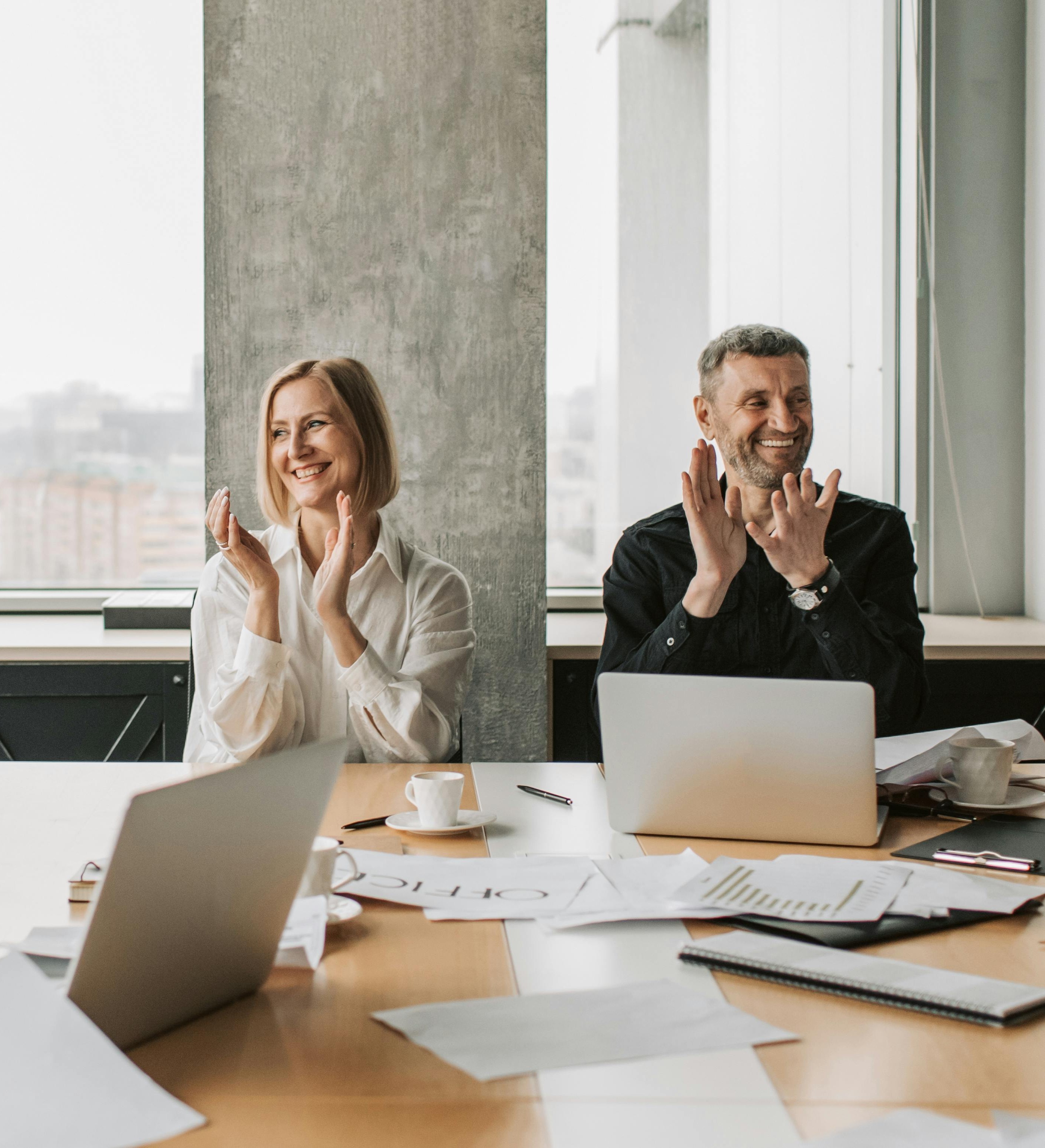 Two colleagues sitting at a conference table with laptops and papers, smiling and clapping hands.