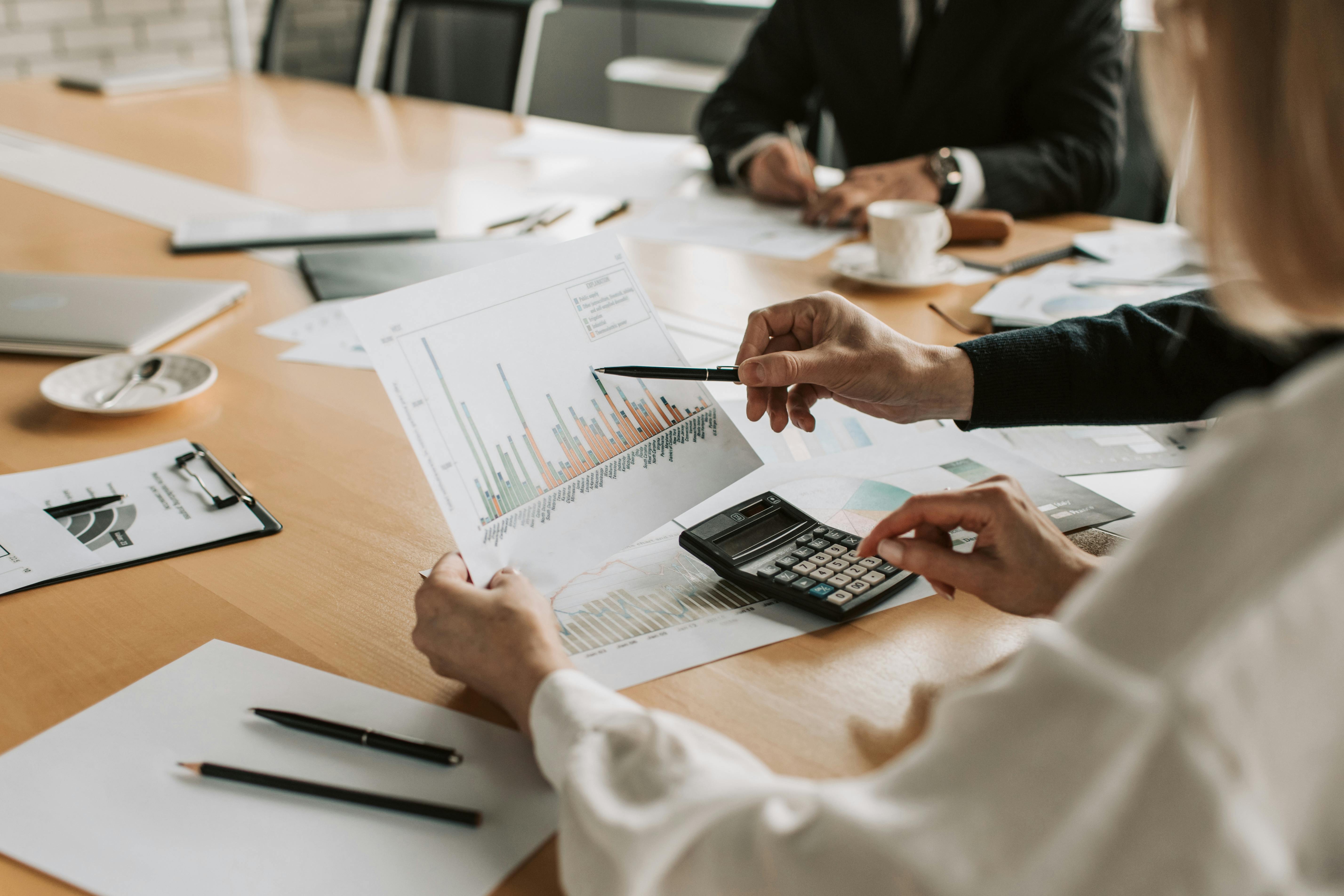 Person using a calculator and pointing at a bar chart on a document during a business meeting.