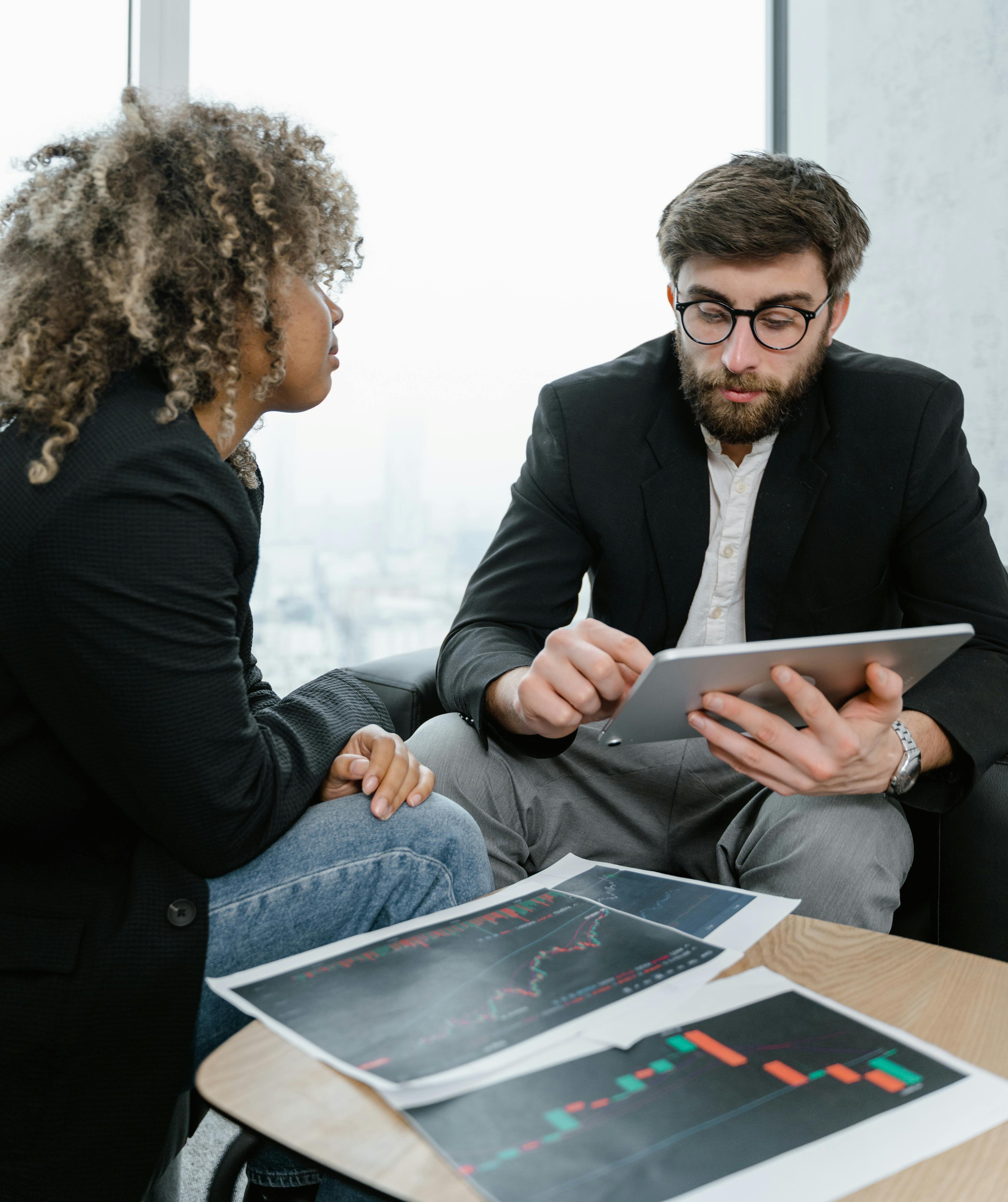 Two colleagues in business attire reviewing financial charts and using a tablet in a modern office.