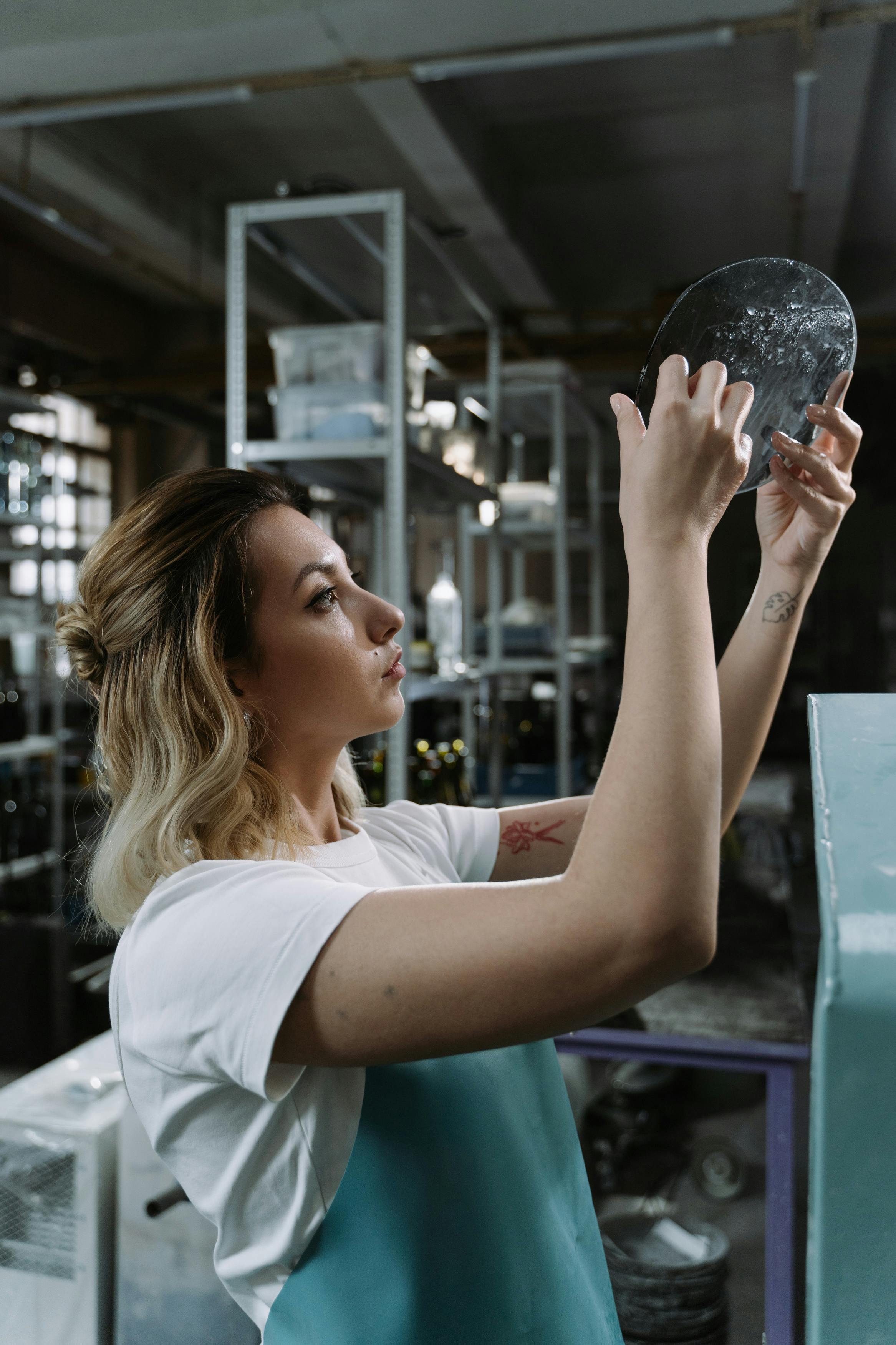 Woman wearing a white shirt and blue apron examining a round black object in a workshop.