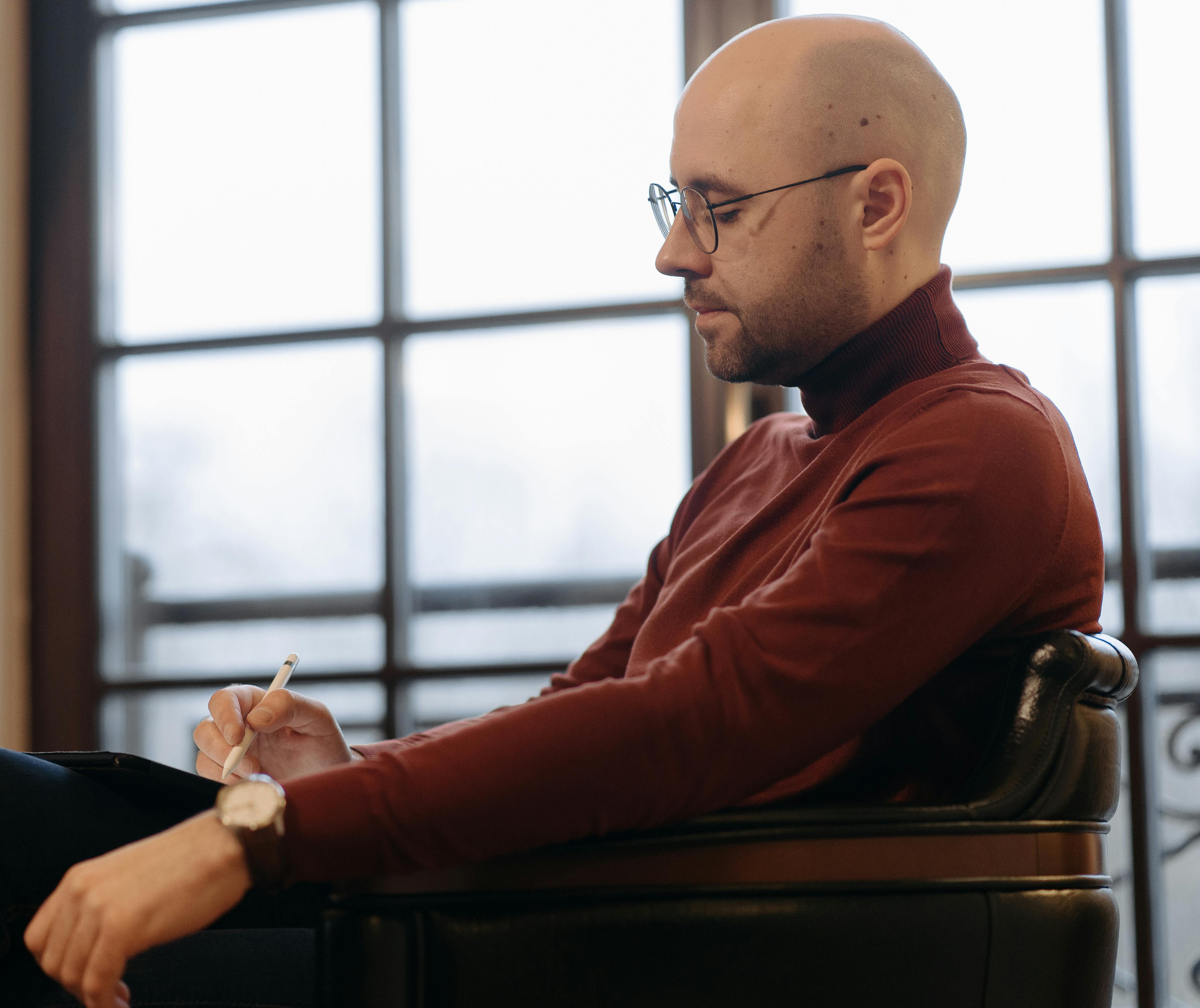 Bald man wearing glasses and brown turtleneck sitting in a chair and writing with a stylus on a tablet in front of a large window.