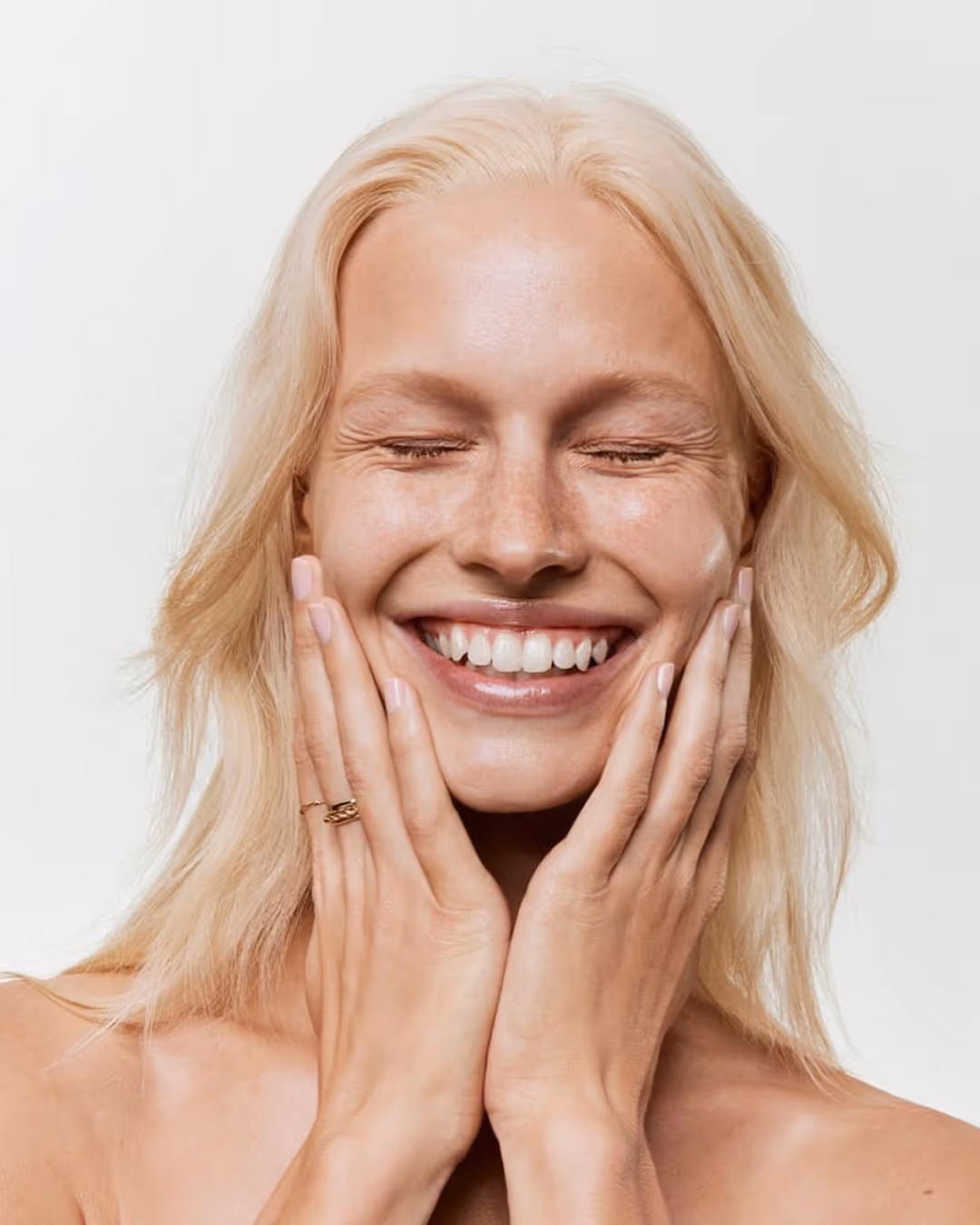 Femme blonde souriante, les yeux fermés et les mains posées sur son visage, exprimant bien-être et légèreté