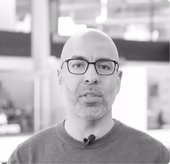 Middle-aged bald man with glasses speaking and gesturing with hands in an indoor setting with blurred background screens.