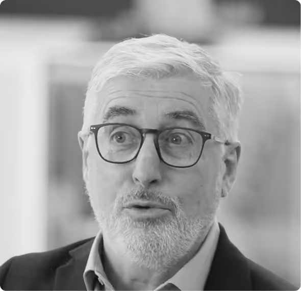 Older man with white hair, beard, and glasses wearing a blazer and shirt, speaking with a microphone attached in an indoor setting.