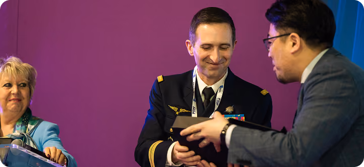 Man in military uniform receiving an award from a man in a suit while a woman watches and smiles.