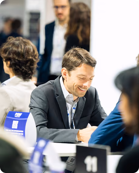Smiling man in black suit sitting at a desk during a busy professional event or meeting.
