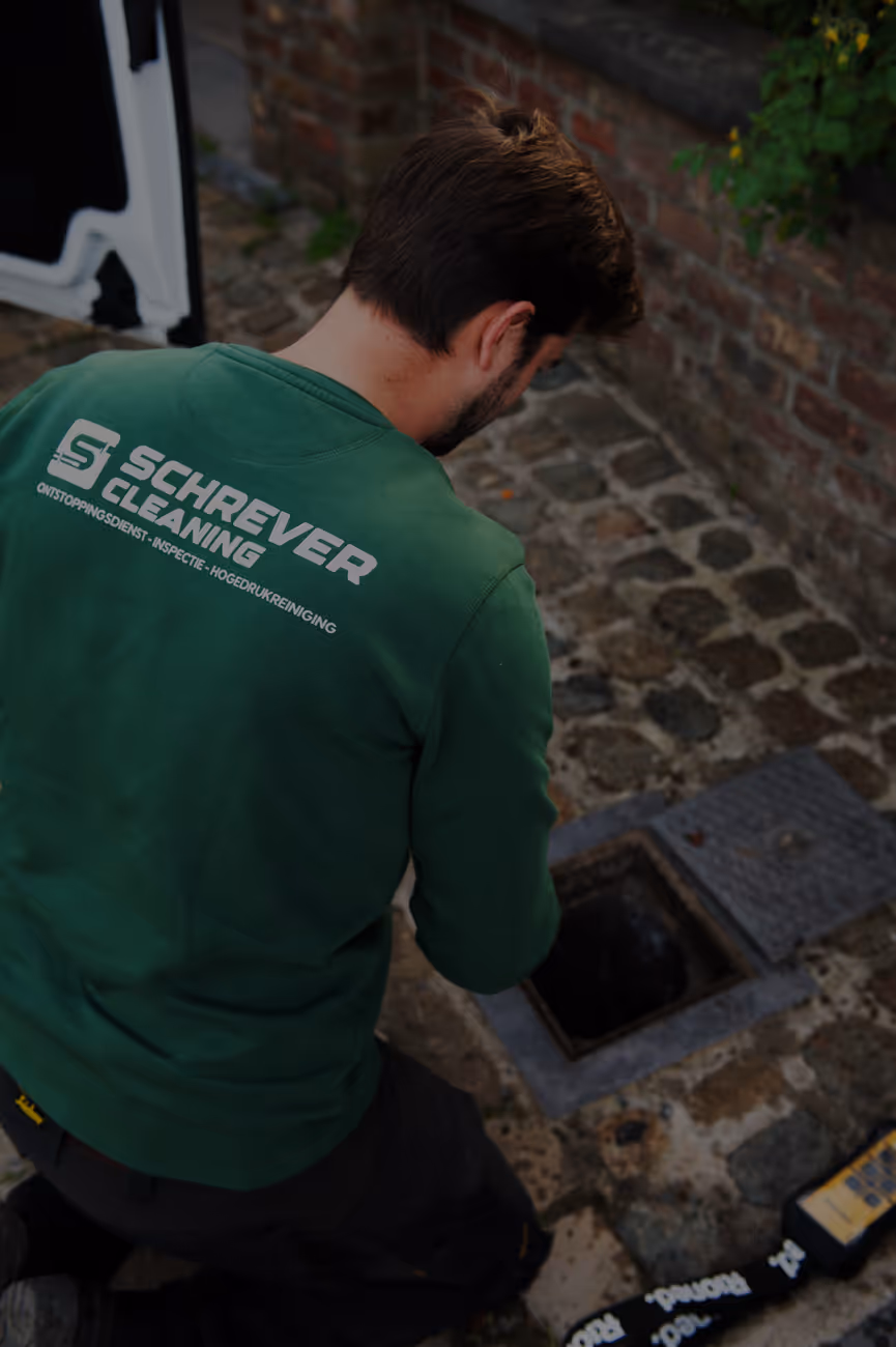Man in een groen shirt met Schrever Cleaning-logo die een put inspecteert op een stenen pad.
