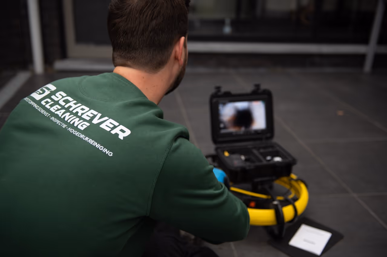 Man met een groene trui met opschrift 'Schrever Cleaning' bestuurt een inspectieapparaat met een monitor en een gele ring op een donkere tegelvloer.