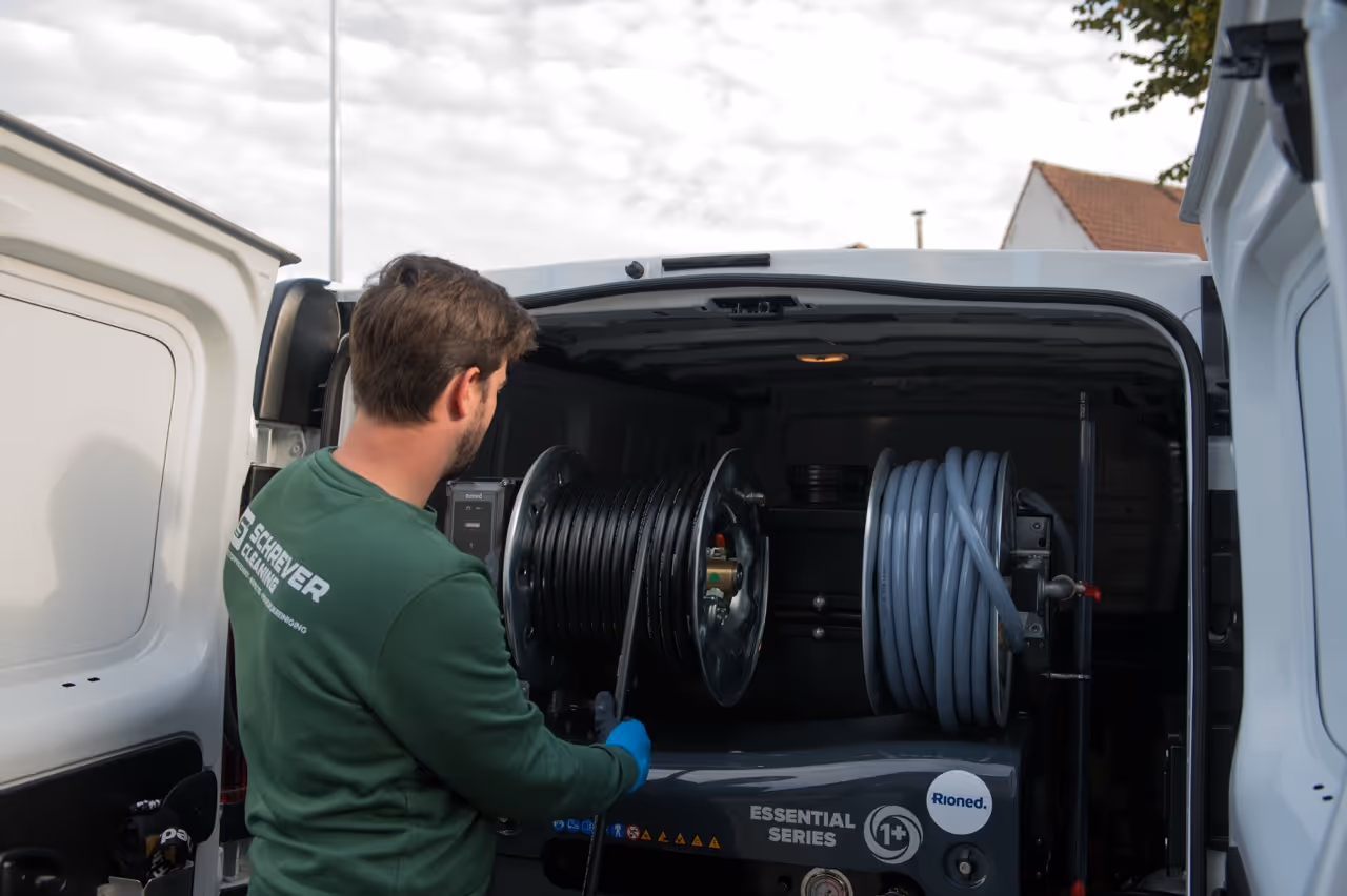 Man met groene trui draait aan haspel met zwarte slang in een witte bedrijfswagen.
