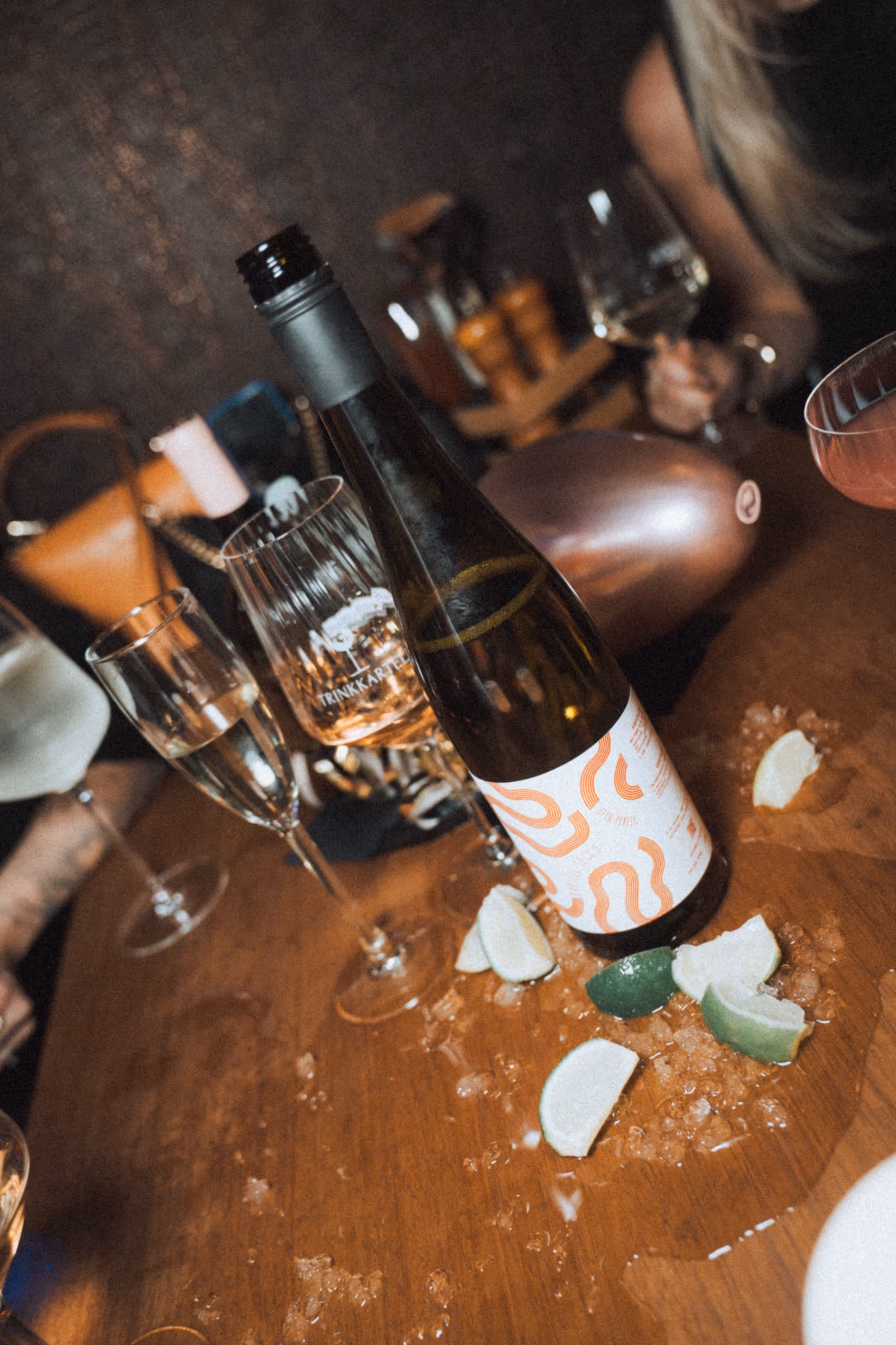Eine Flasche Wein steht auf einem Tisch, um die Flasche herum ist eine Wasserlache, in der Eisstücke und Limettenstücke liegen. Auf dem Tisch sind ebenfalls verschiedene Gläser zu sehen.
