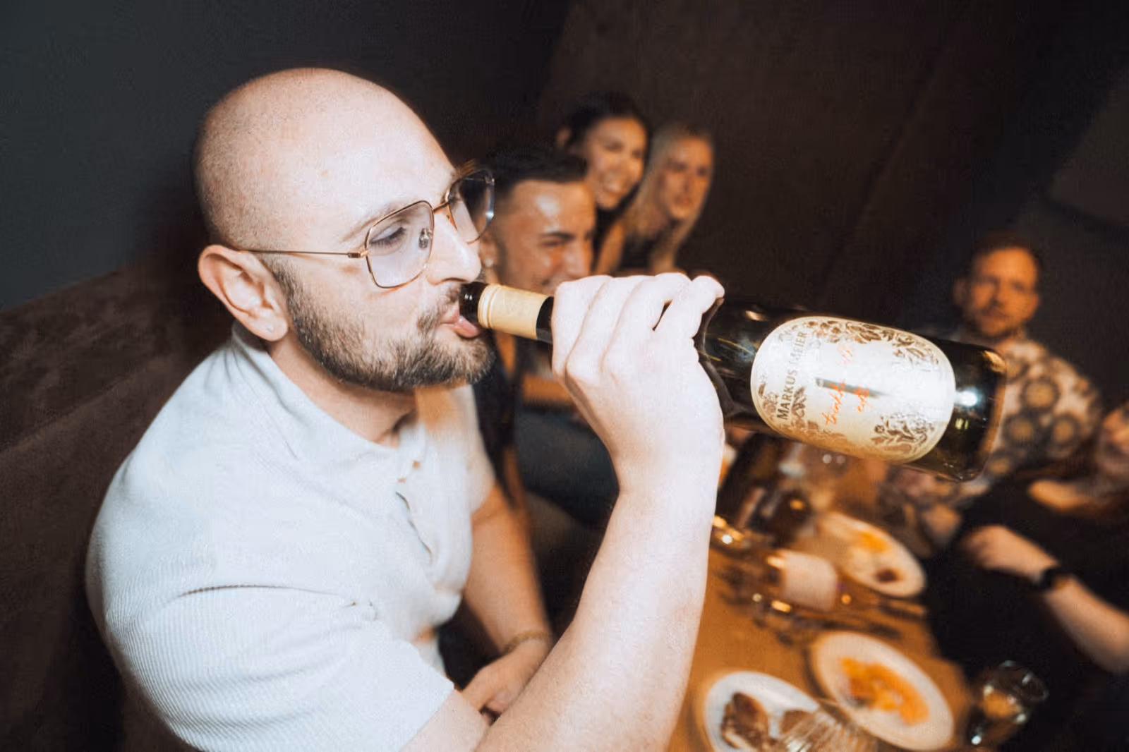 Ein Mann mit Brille trinkt direkt aus einer Weinflasche. Im Hintergrund sind weitere Personen zu sehen, die um einen Tisch herum sitzen und lachen.