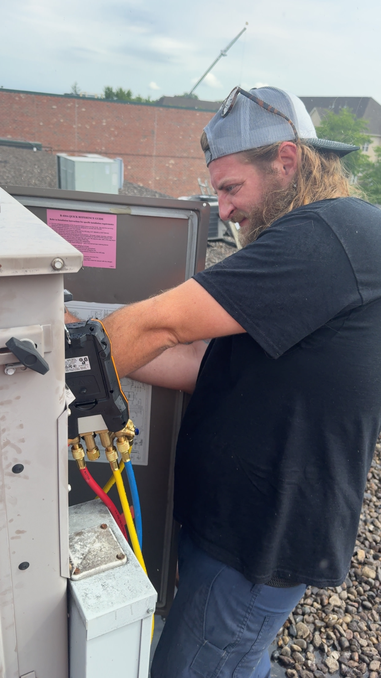 A technician checks and maintains an HVAC unit using colorful service hoses on a rooftop.

