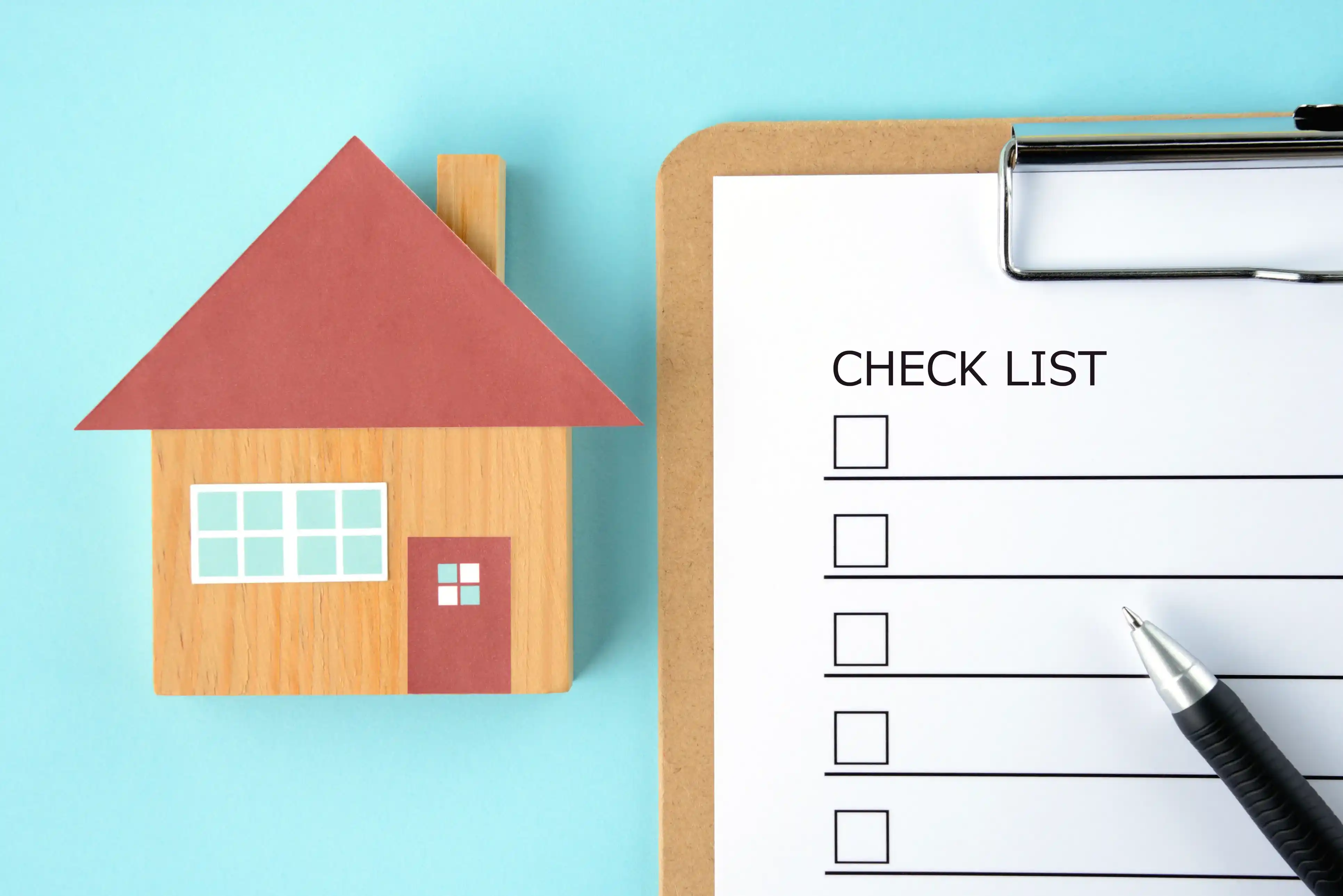 Overhead view of a wooden miniature house next to a clipboard labeled "CHECK LIST" with a pen, on a blue background.