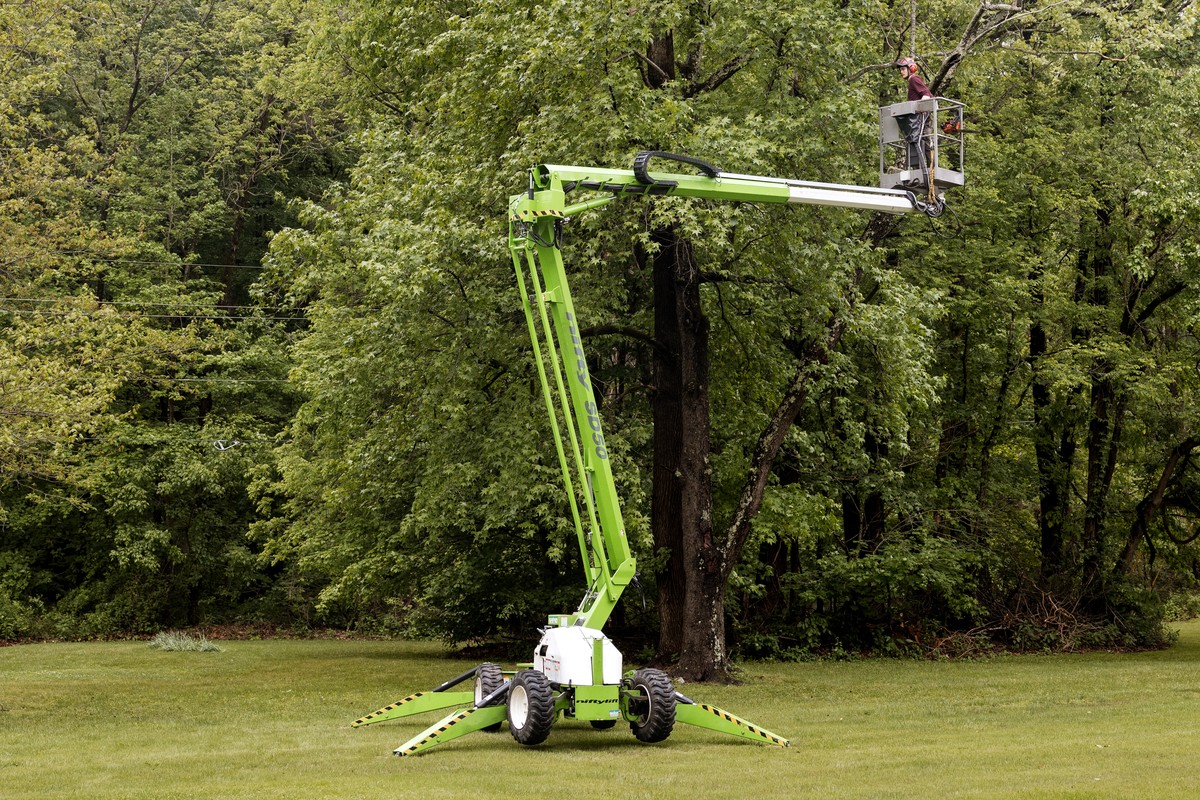 Tree-service worker trimming branches from an aerial lift surrounded by green trees.