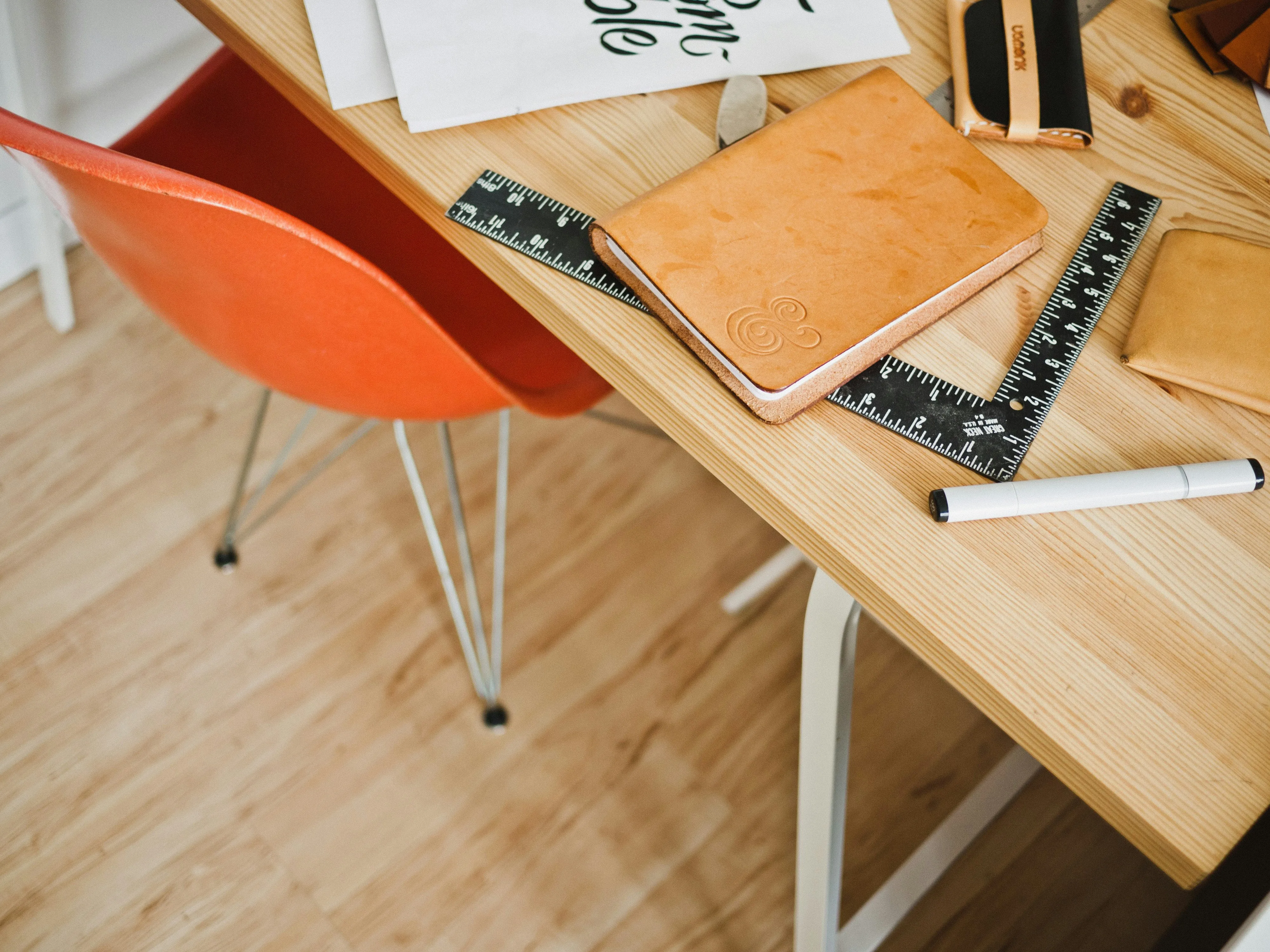 A top-down zoomed in picture of a pine-colored wooden desk with white metal legs, an orange molded plastic chair with white metal legs pushed into the desk at a slight angle; on the desk is a black L-shaped ruler, a leather-bound notebook, a used-white rectangular eraser, and some blank sheets of paper with some illegible black calligraphy showing