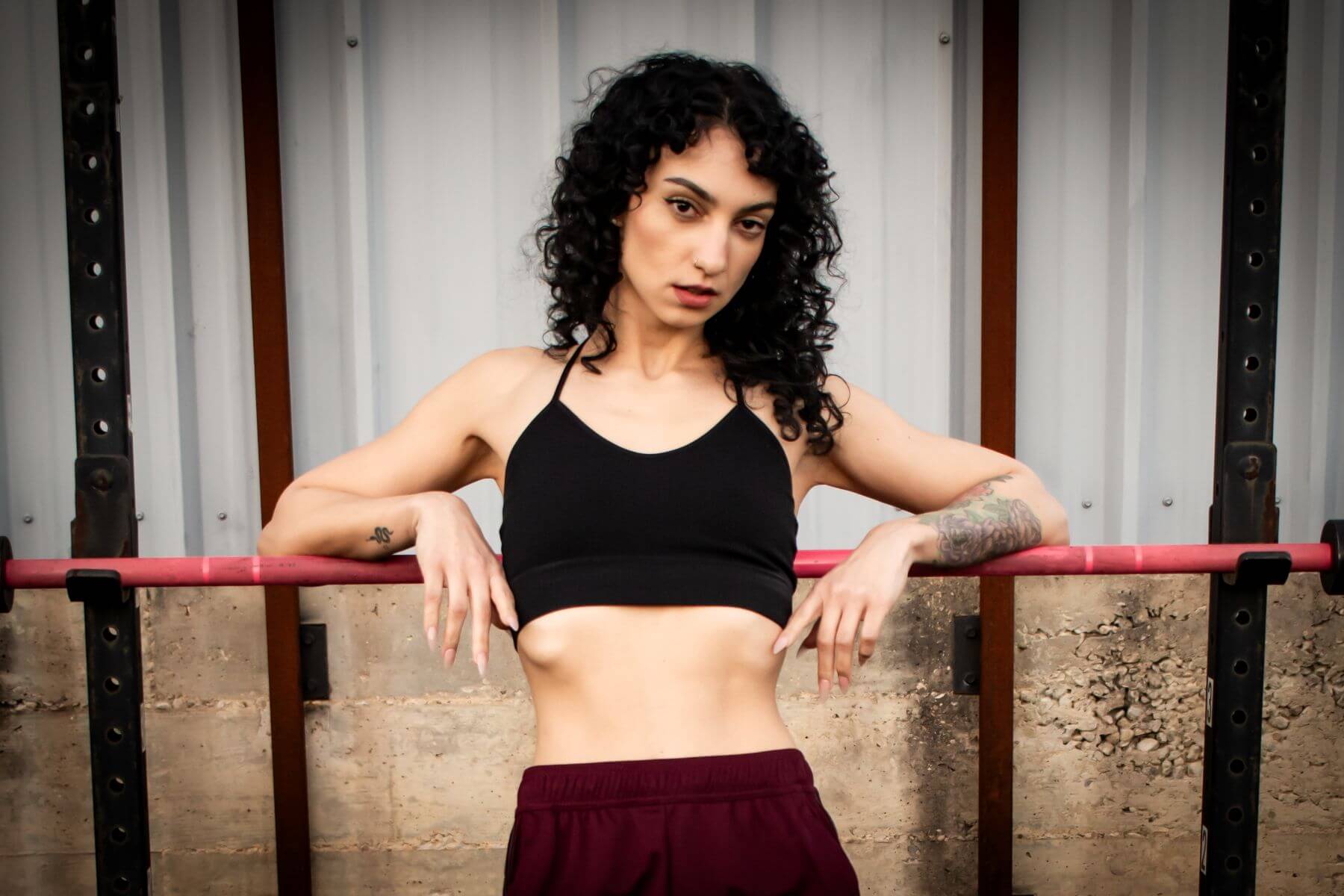 Young woman in athletic attire leaning on barbell of a squat rack in a gym.