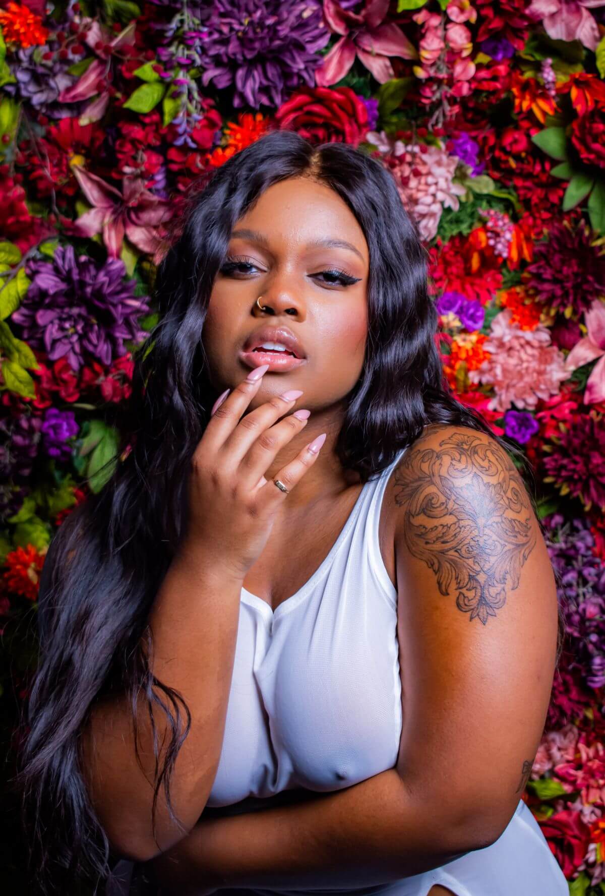 Woman in thin white dress, fingers to her lip, standing against a vibrant wall of flowers.