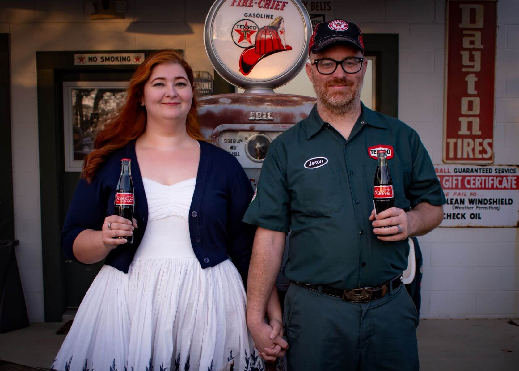 Couple holding hands at retro gas station, sharing a coke. Woman in white dress, man in retro mechanic wear.