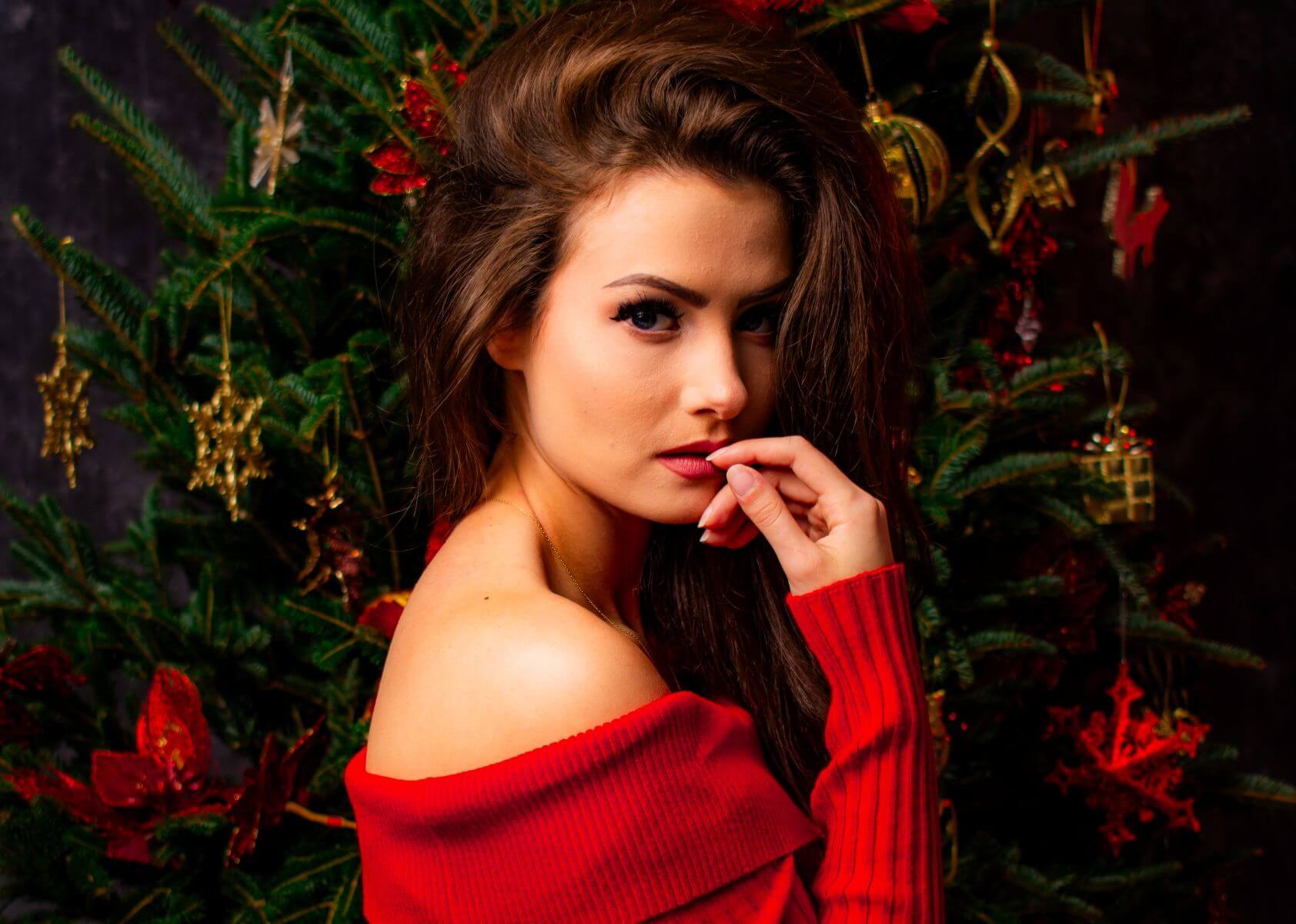 Young brunette posing in off shoulder red sweater, finger to red lips, in front of Christmas tree.