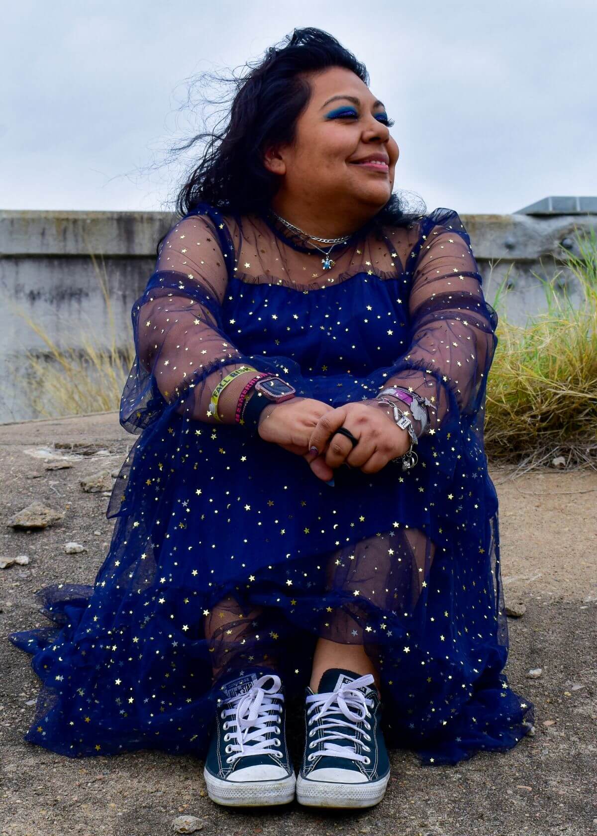 Woman in blue dress and Converse, sitting in front of lake, knees to chest.
