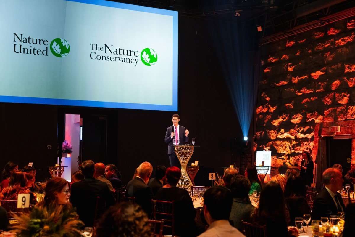 A man in a suit speaks at a podium during a formal indoor event with seated guests and Nature United and The Nature Conservancy logos projected on a screen.