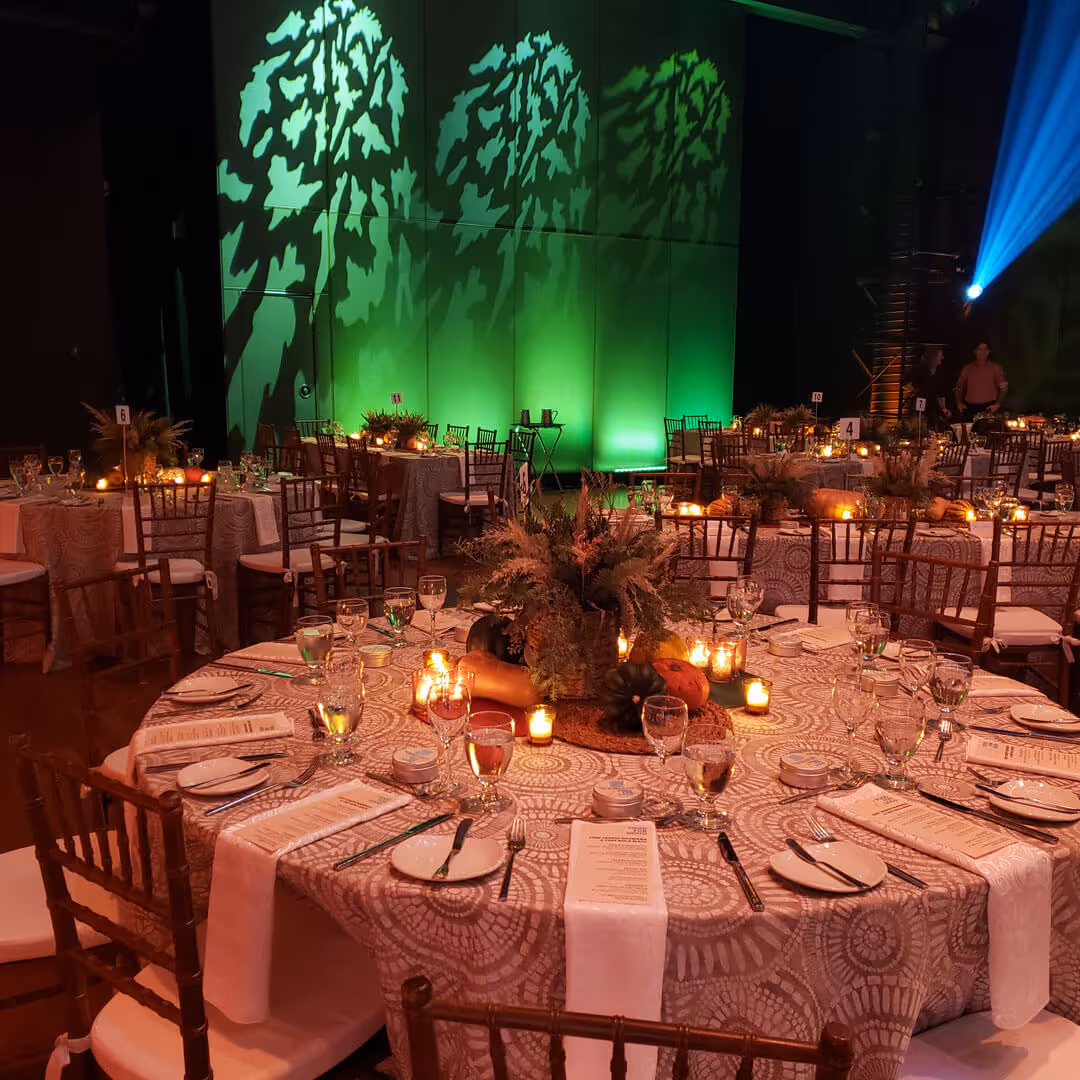Round tables set for a formal dinner with patterned tablecloths, floral centerpieces, candles, and green tree-shaped light projections on a wall in the background.
