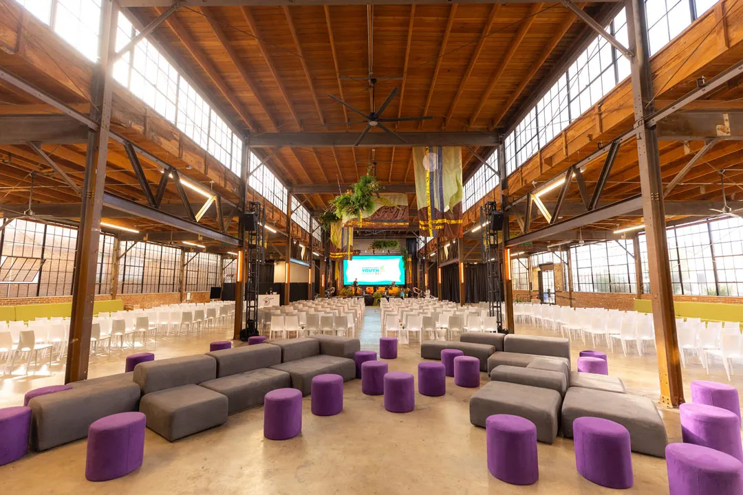 Spacious industrial-style event hall with rows of white chairs facing a stage, surrounded by gray modular sofas and purple ottomans.