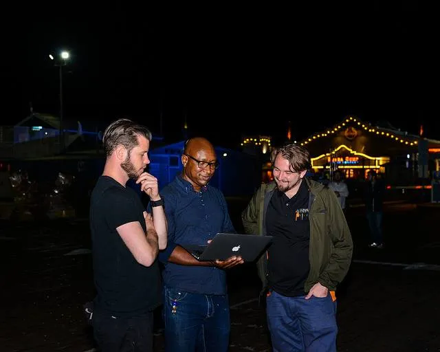 Three men standing outdoors at night, one holding a laptop while the others look on attentively.