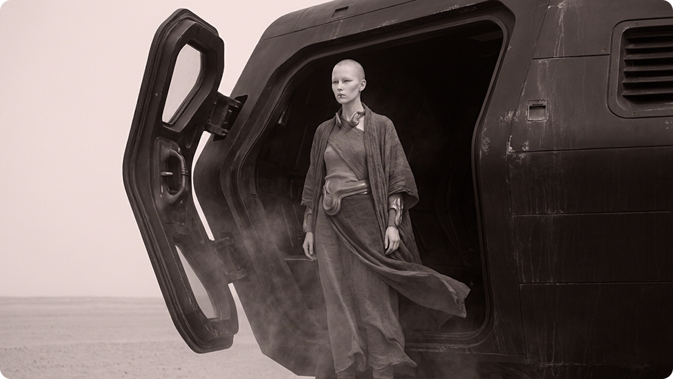Bald person in flowing, futuristic robes standing at the open hatch of a large dark vehicle in a misty desert landscape.