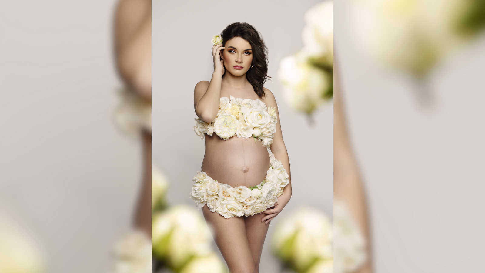 Donna incinta in posa, adornata con composizioni floreali di rose bianche e color crema che coprono il busto e il corpo inferiore, formando un abito decorativo. Un fiore singolo è posizionato tra i capelli. Lo sfondo sfocato con elementi floreali simili mette in risalto il soggetto centrale. Lo stile artistico celebra la gravidanza, la femminilità e la connessione con la natura.