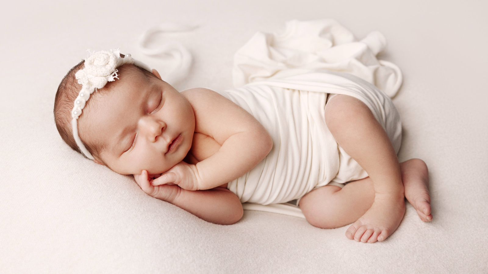 Neonato che dorme serenamente su una superficie morbida e chiara, avvolto in un telo bianco. È sdraiato su un fianco con le mani vicino al viso. Indossa una delicata fascia con decorazioni floreali sulla testa. L'immagine trasmette un senso di innocenza, tranquillità e tenerezza.