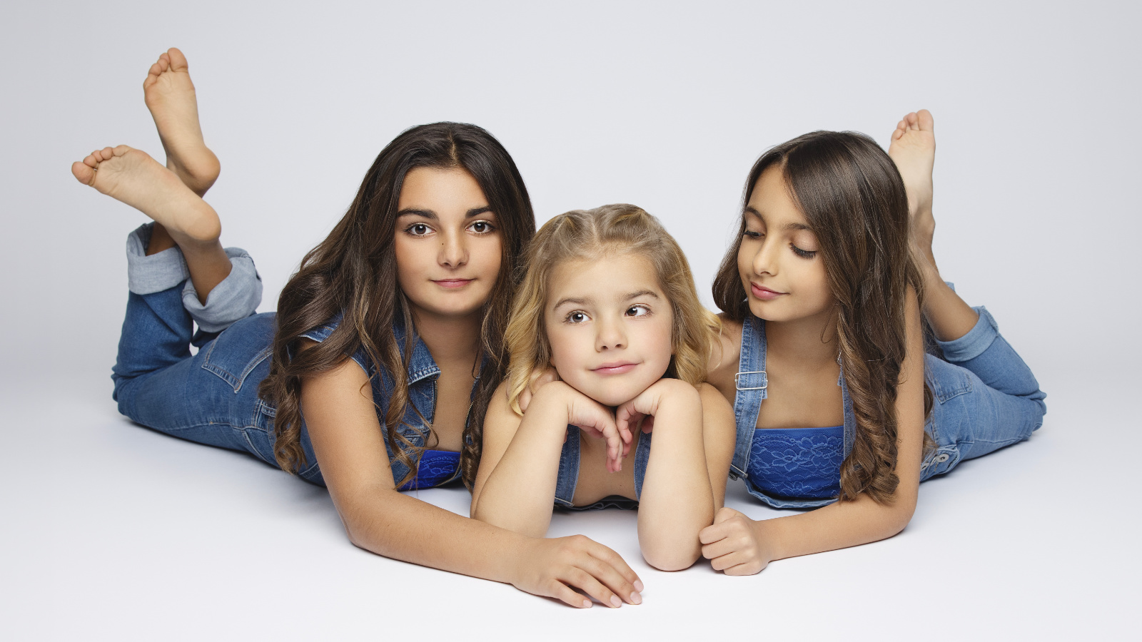 Tre bambini con capelli lunghi e abiti blu sono sdraiati sul pavimento bianco. Il bambino al centro sostiene il mento con le mani, mentre gli altri due hanno le gambe sollevate e incrociate. L'immagine trasmette simmetria, naturalezza e complicità.
