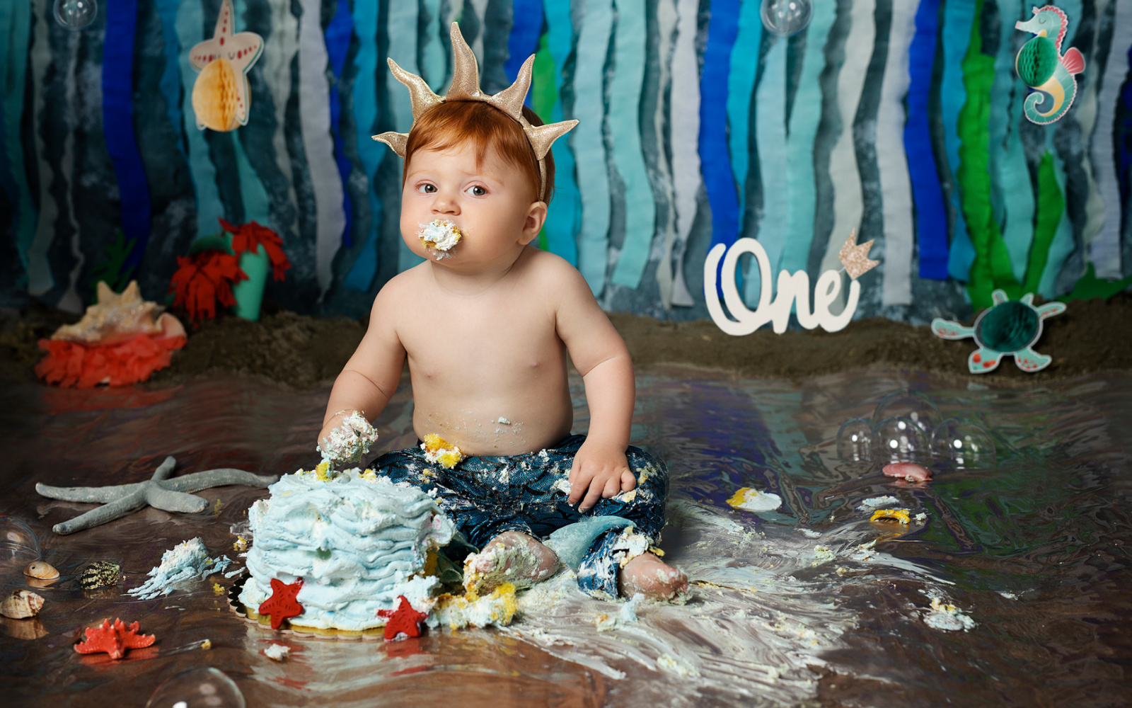 Bambino con corona corallo e torta blu distrutta in servizio fotografico smash cake primo compleanno a tema La Sirenetta.