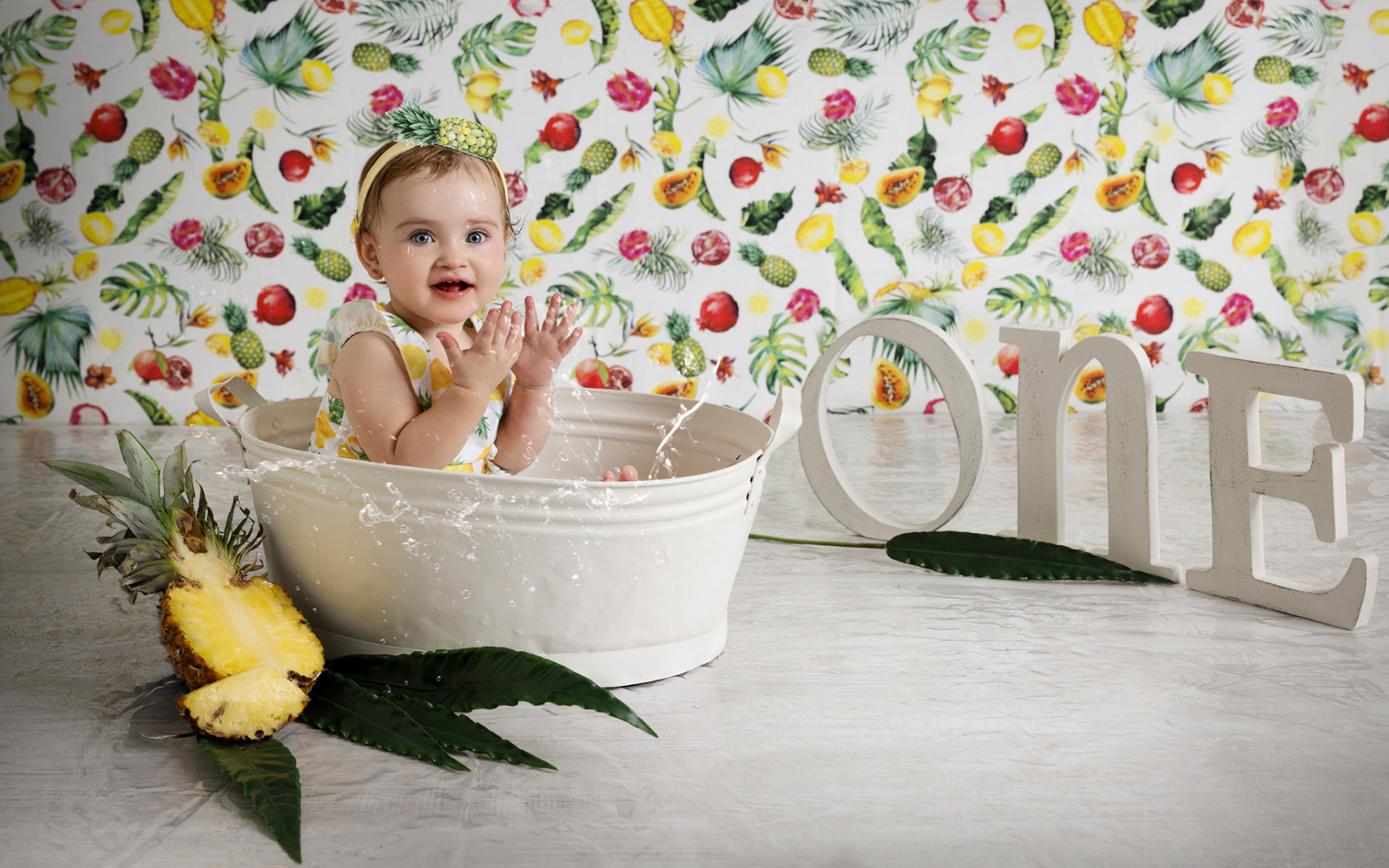 Bambina in tinozza bianca con outfit giallo e sfondo frutta tropicale in servizio fotografico smash cake primo compleanno.