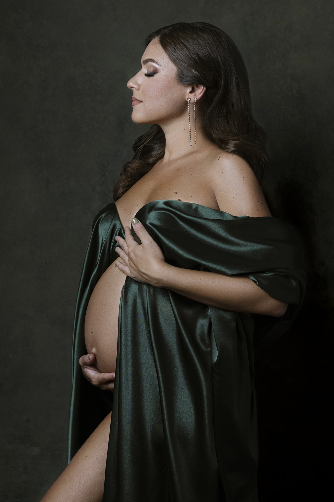 Servizio fotografico maternità a Bologna con Claudia Spaziani: donna incinta in satin verde scuro, posa laterale e luce morbida, atmosfera raffinata.