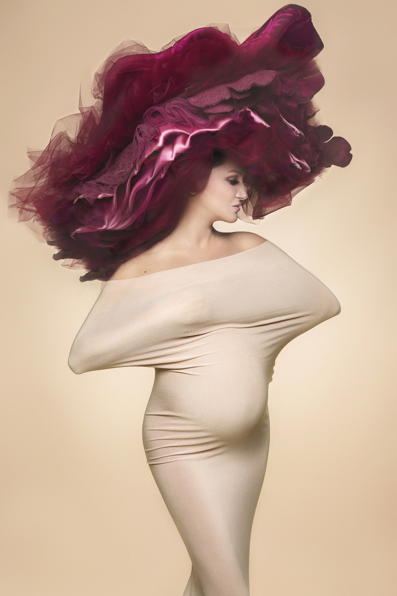 Foto maternità fashion a Bologna con Claudia Spaziani: donna incinta in outfit beige scultoreo e copricapo in tulle bordeaux, stile concettuale e d’impatto.