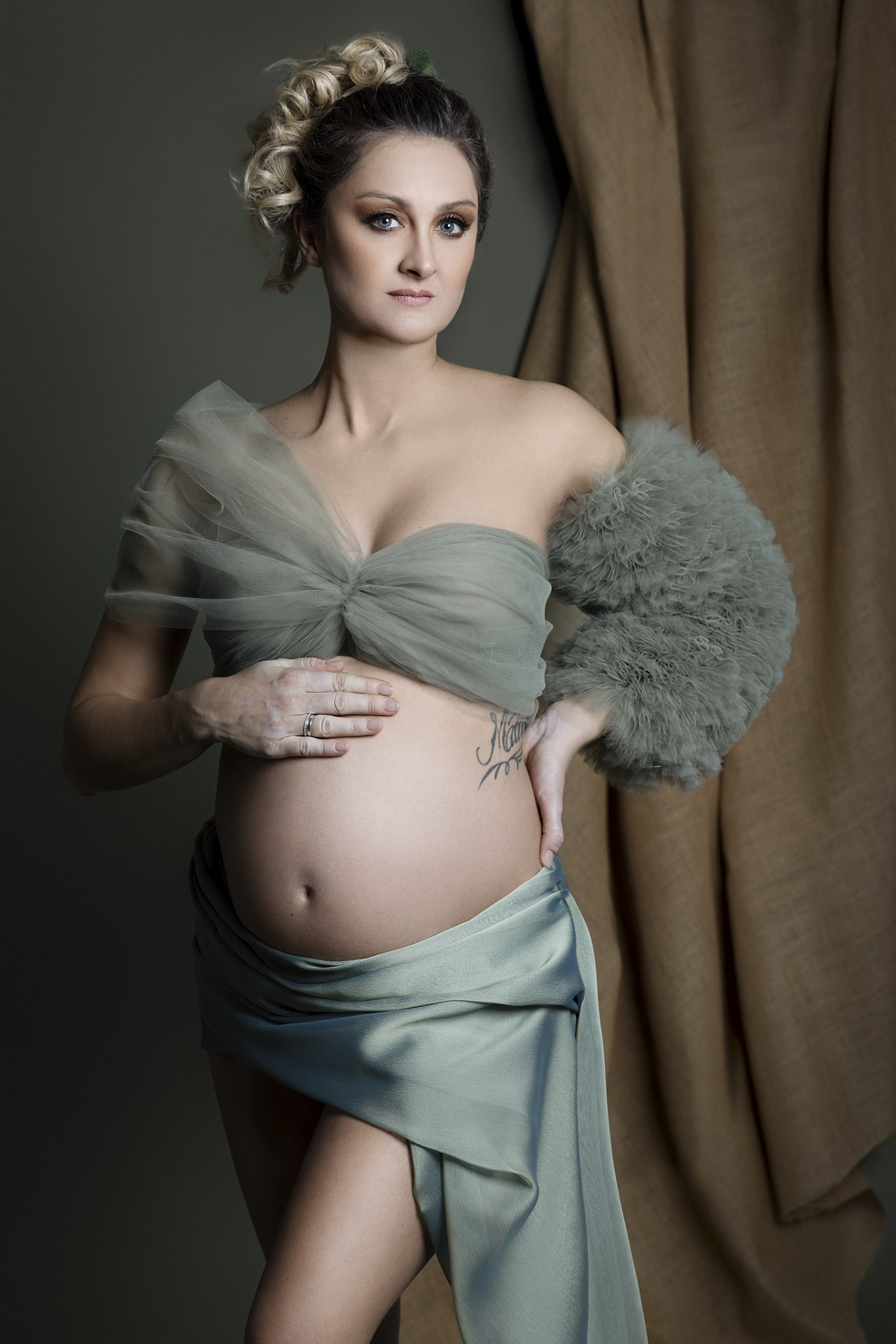 Foto maternità a Bologna firmata Claudia Spaziani: donna incinta avvolta in tessuto verde salvia trasparente, dettaglio tattoo e atmosfera raffinata.