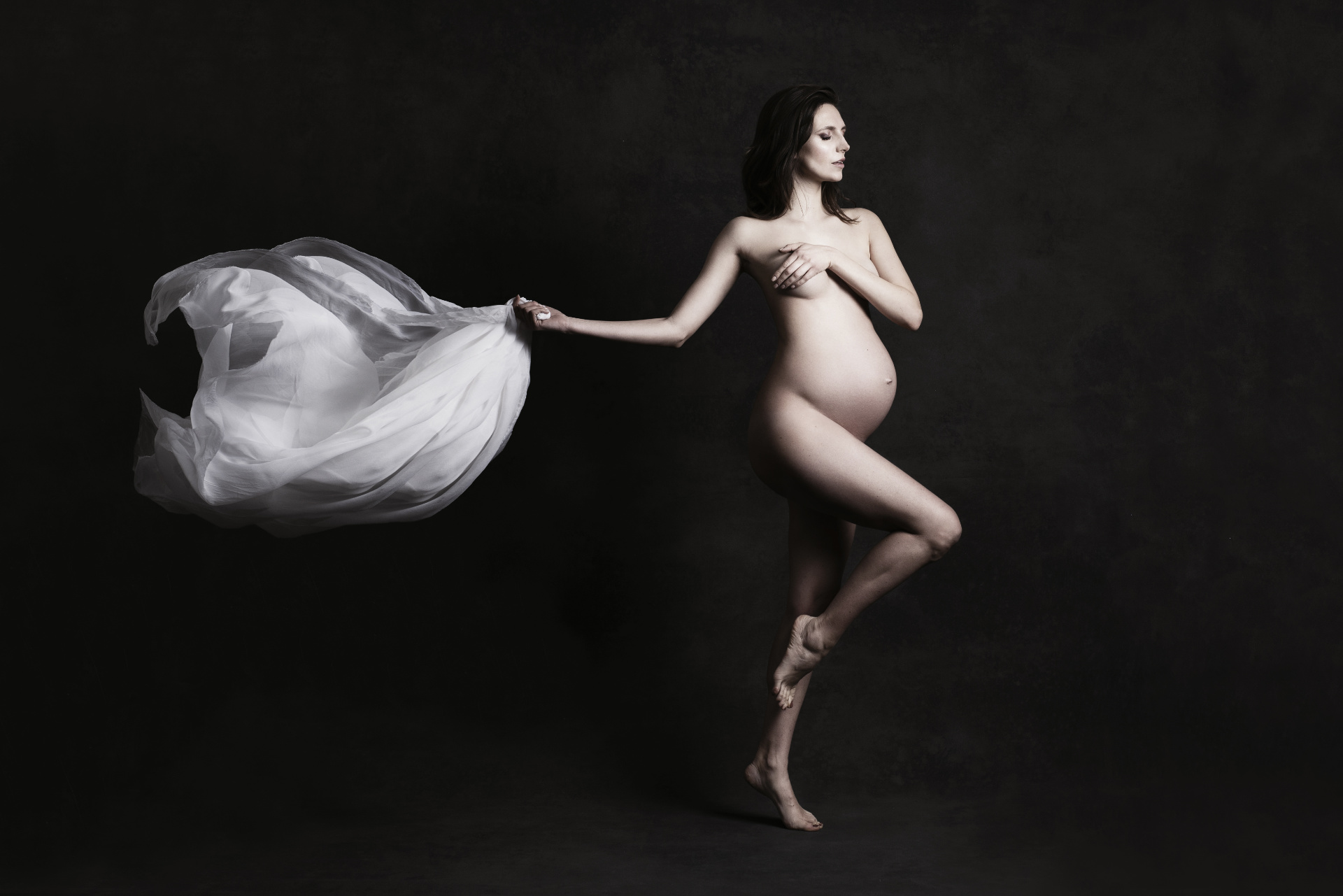 Foto artistica di maternità a Bologna realizzata da Claudia Spaziani: donna incinta in posa elegante con tessuto bianco fluente, ispirazione danza e luce drammatica.