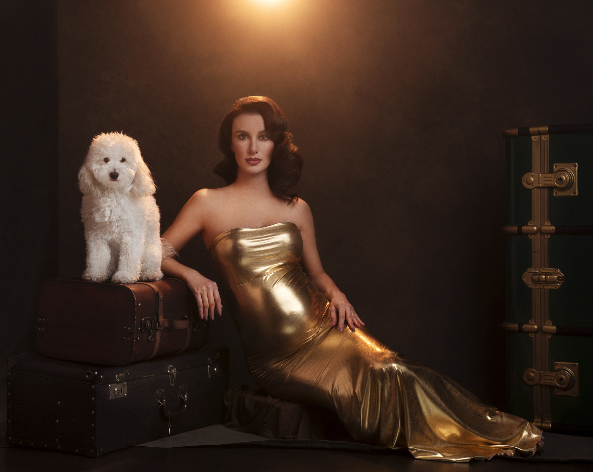 Servizio fotografico maternità glamour a Bologna con Claudia Spaziani: donna incinta in abito dorato accanto a valigie vintage e cane bianco, atmosfera sofisticata.