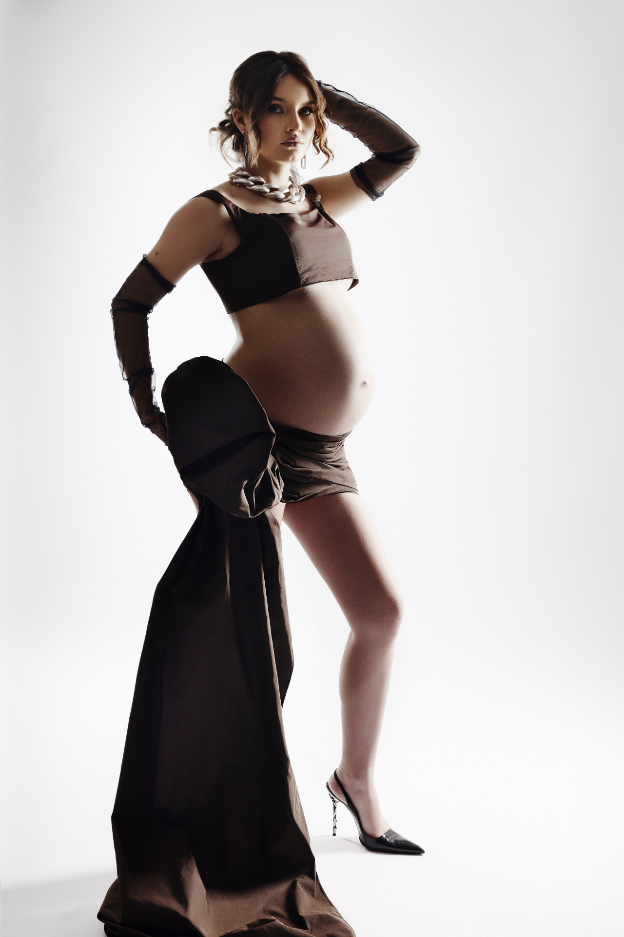 Servizio fotografico maternità a Bologna con Claudia Spaziani: donna incinta in outfit marrone con top corto, guanti lunghi e gioielli, posa fashion e decisa.
