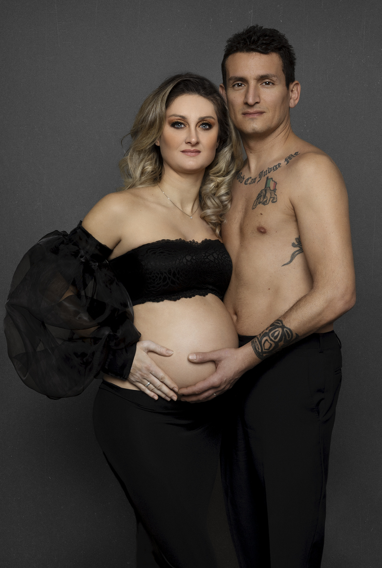 Servizio fotografico maternità a Bologna con Claudia Spaziani: coppia in posa intima, donna incinta in top nero e partner tatuato, atmosfera emozionale.