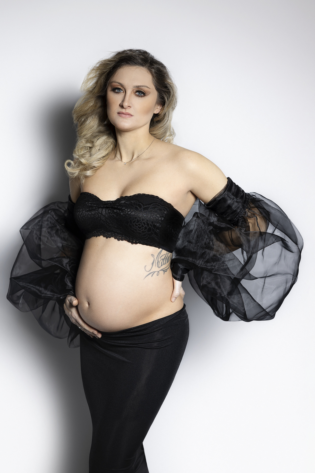 Ritratto di maternità a Bologna realizzato da Claudia Spaziani: donna incinta in top di pizzo nero e lunga gonna, tatuaggio “Mateo” visibile, stile elegante.