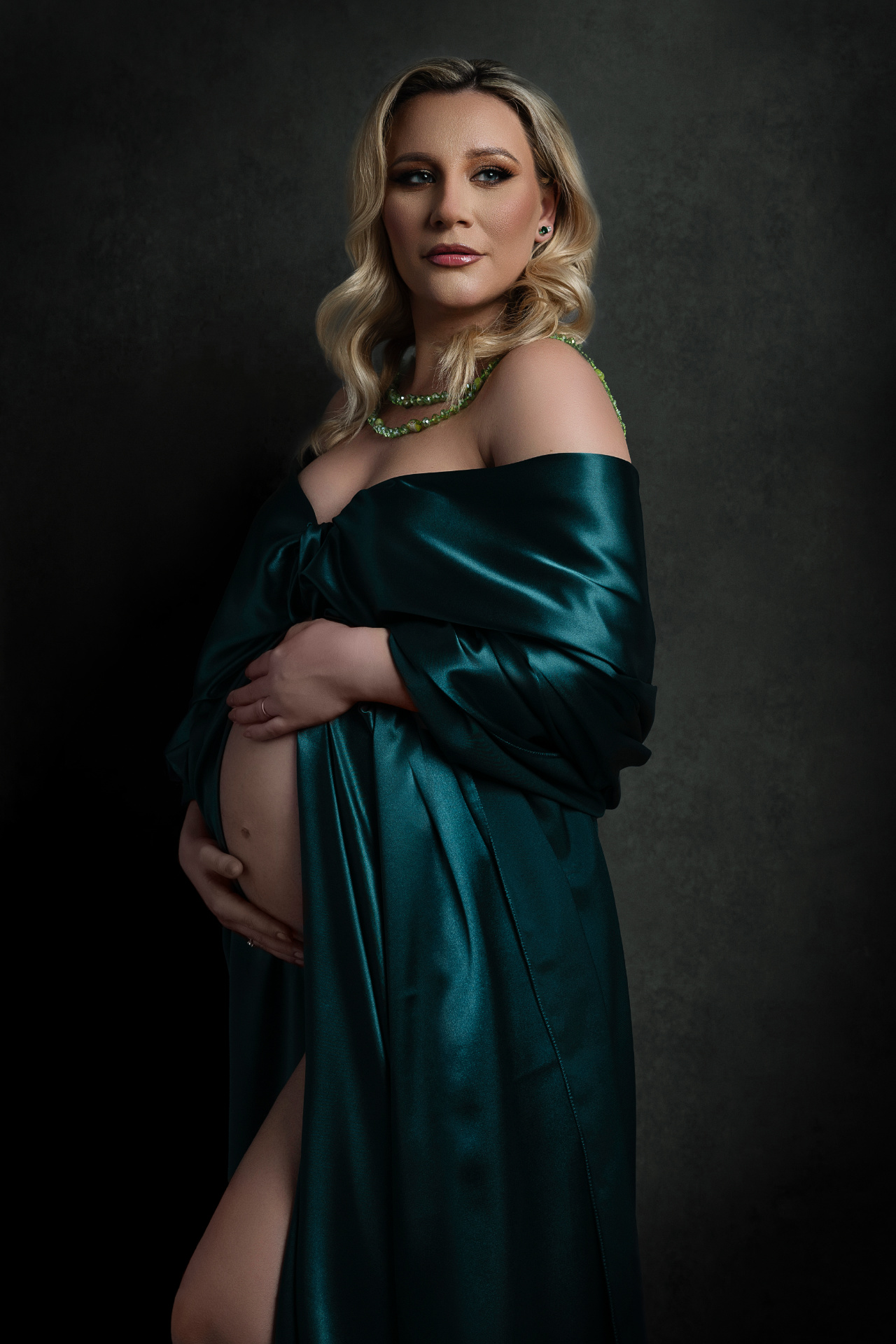 Foto maternità a Bologna firmata Claudia Spaziani: donna incinta in abito satin verde petrolio con collana di perle verdi, luce drammatica e raffinata.