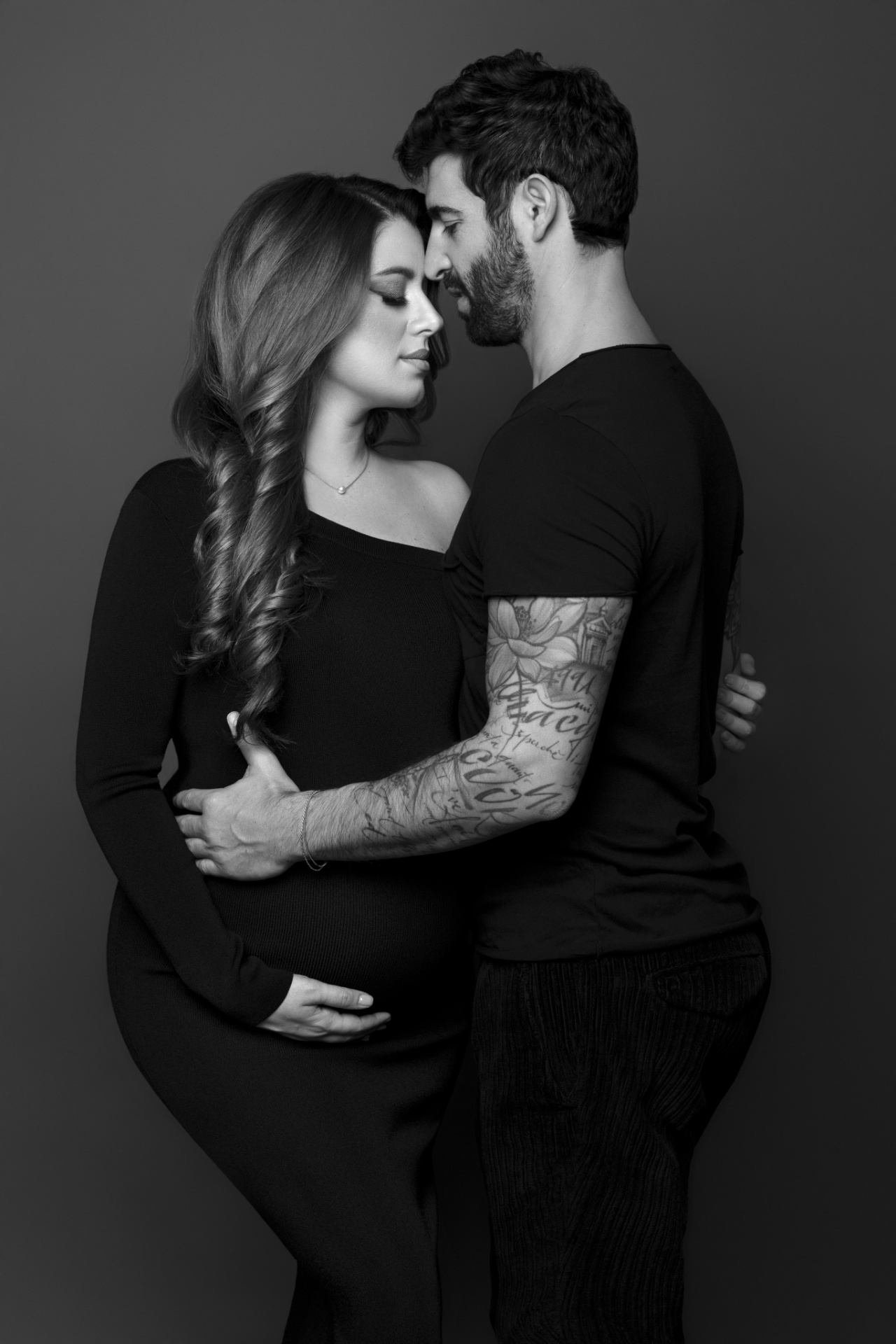 Ritratto di coppia maternità in bianco e nero a Bologna realizzato da Claudia Spaziani: donna incinta in abito nero e partner tatuato, posa intima e affettuosa.