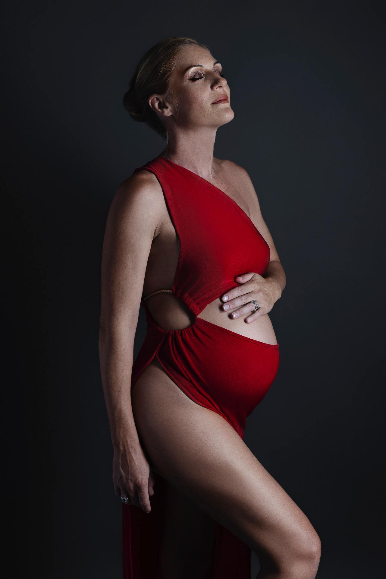 Foto maternità a Bologna con Claudia Spaziani: donna incinta in abito rosso asimmetrico con spacco, posa laterale e luce drammatica.